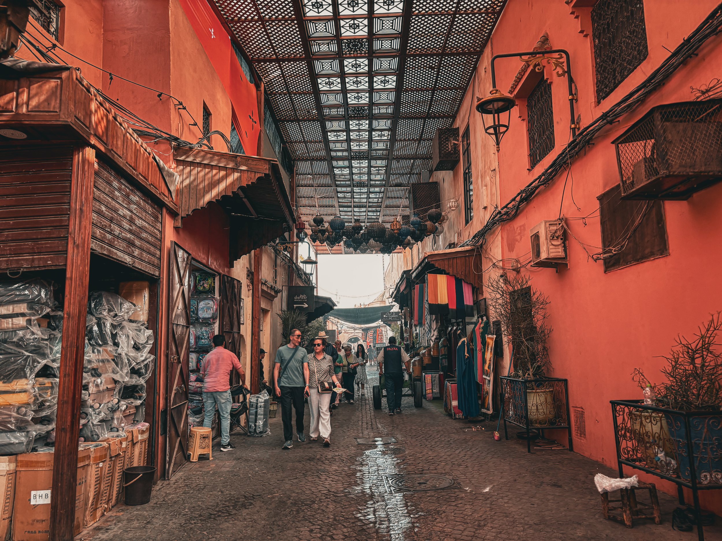 A bustling marketplace with people walking along a cobblestone street under a decorative metal roof. The street is lined with shops on both sides, selling various goods, including textiles and souvenirs. The walls are painted in warm earthy tones, and hanging lanterns add to the vibrant atmosphere.
