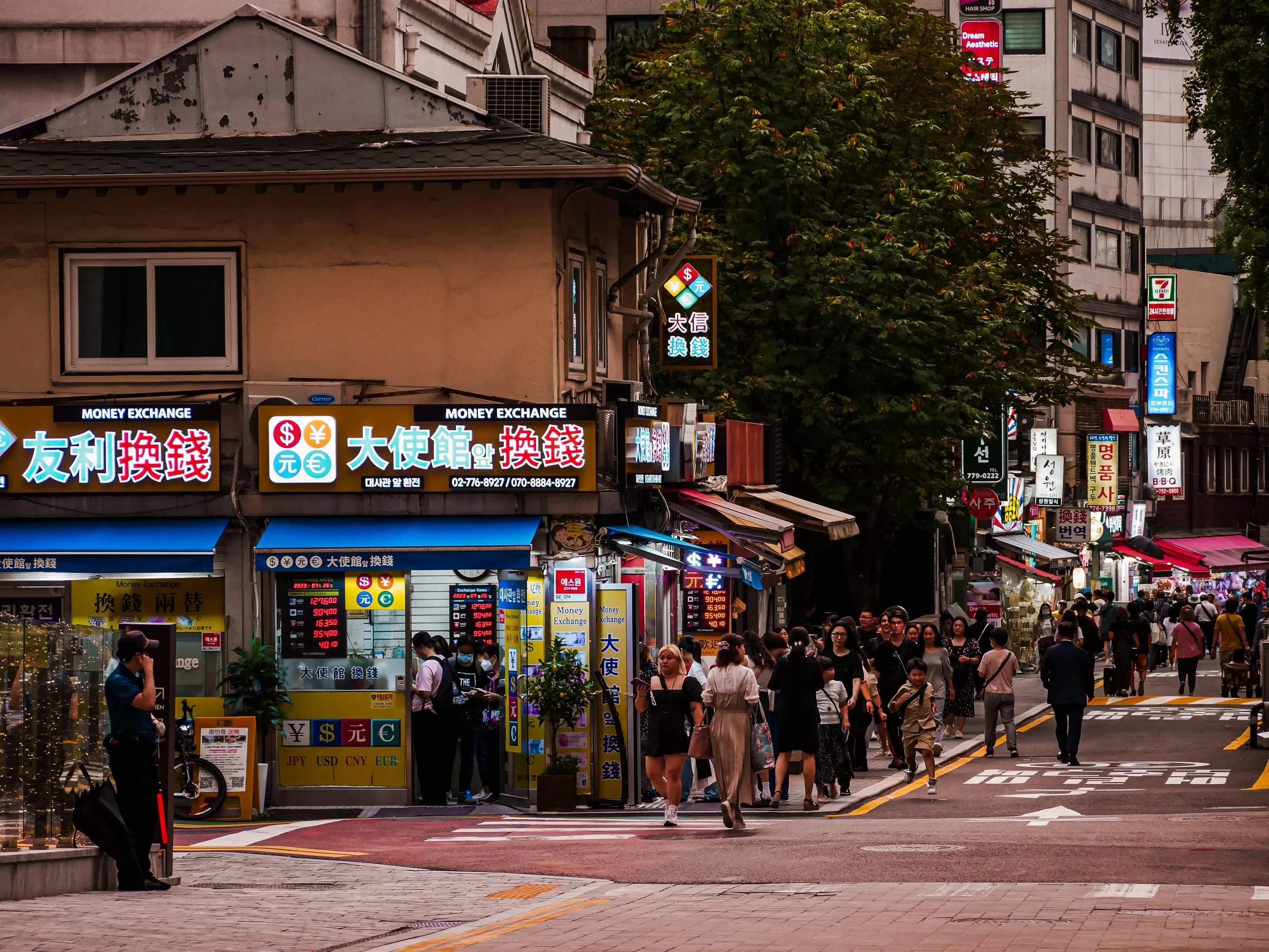 A busy urban street scene filled with pedestrians walking and shopping small storefronts with illuminated signs, including currency exchange services, and tall buildings in the background. There are trees along the sidewalk and a crosswalk in the foreground.
