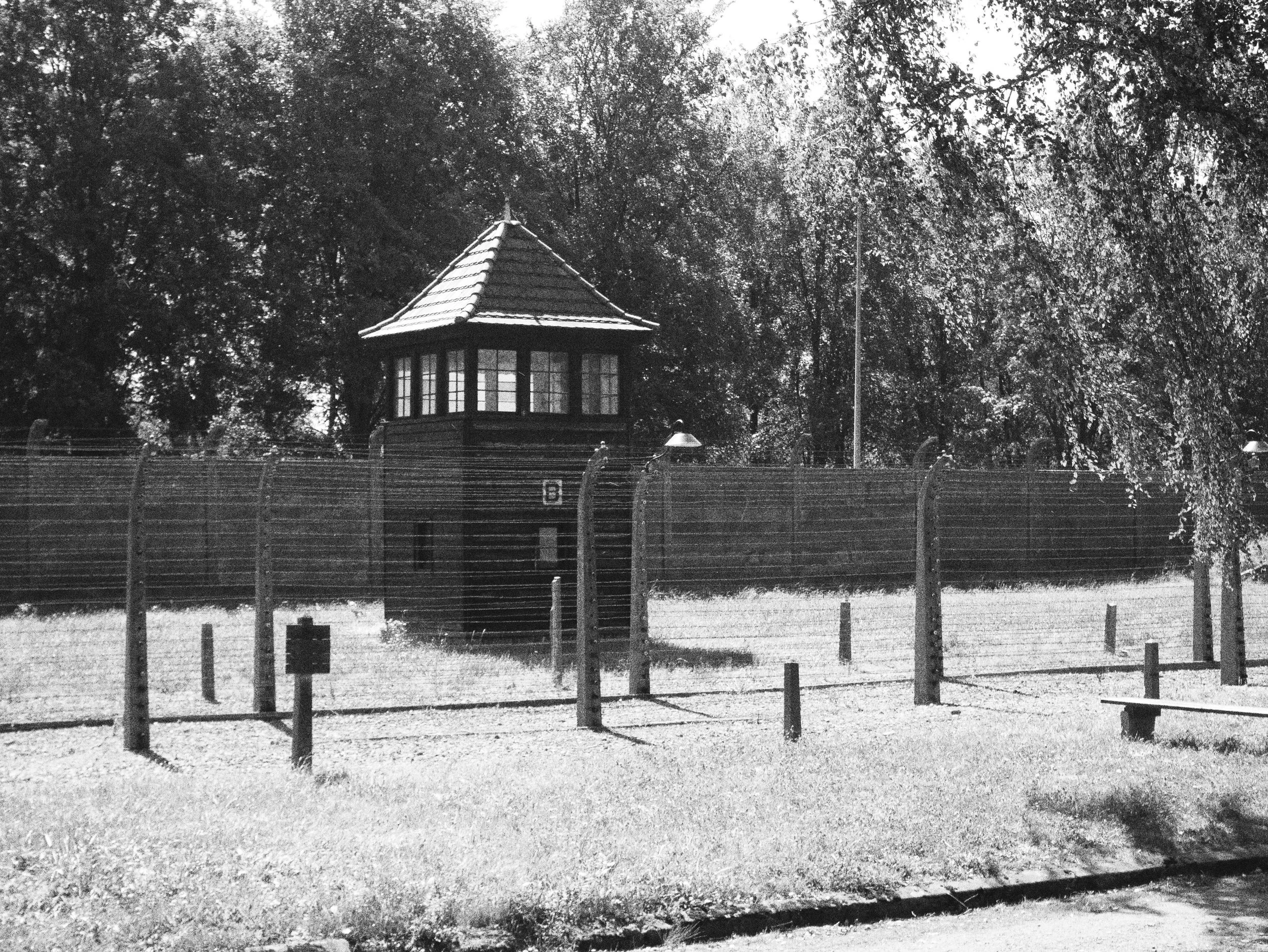 A black and white photo of a small, wooden, pointed-roof guard tower surrounded by a tall fence with barbed wire, set among trees.