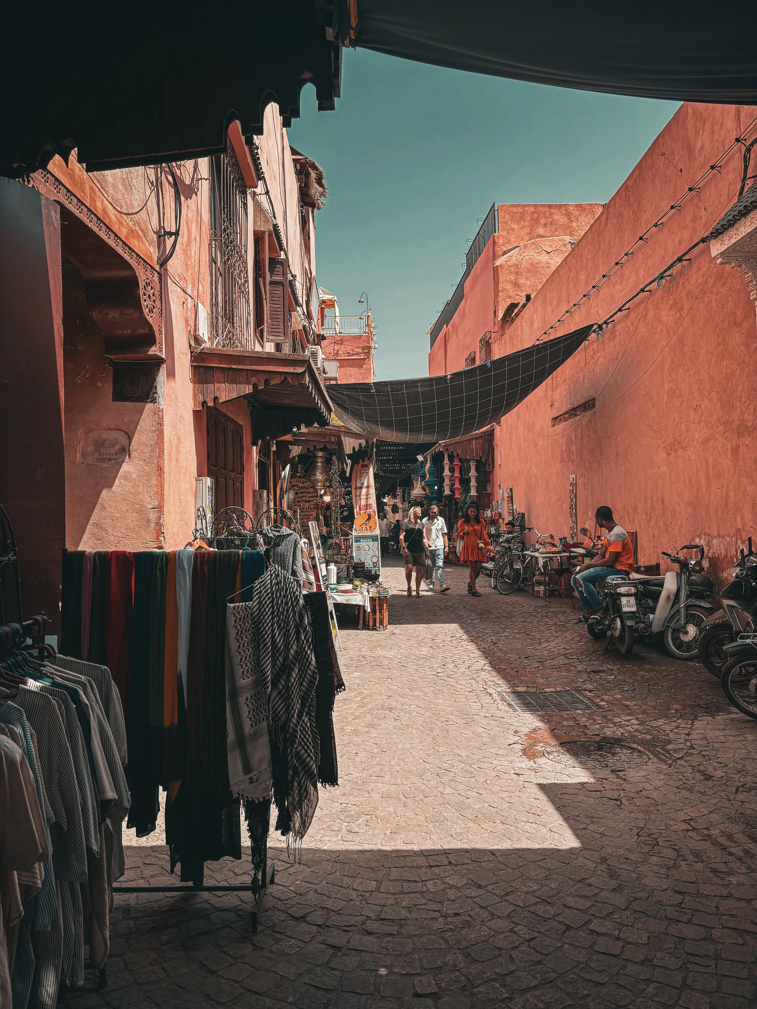 A narrow street with vendors on both sides, colorful clothing hanging on racks, and people walking or sitting on scooters. Old buildings with a mix of wooden and stucco facades line the street, and a black canopy extends over part of the walkway. The sky is clear and blue.