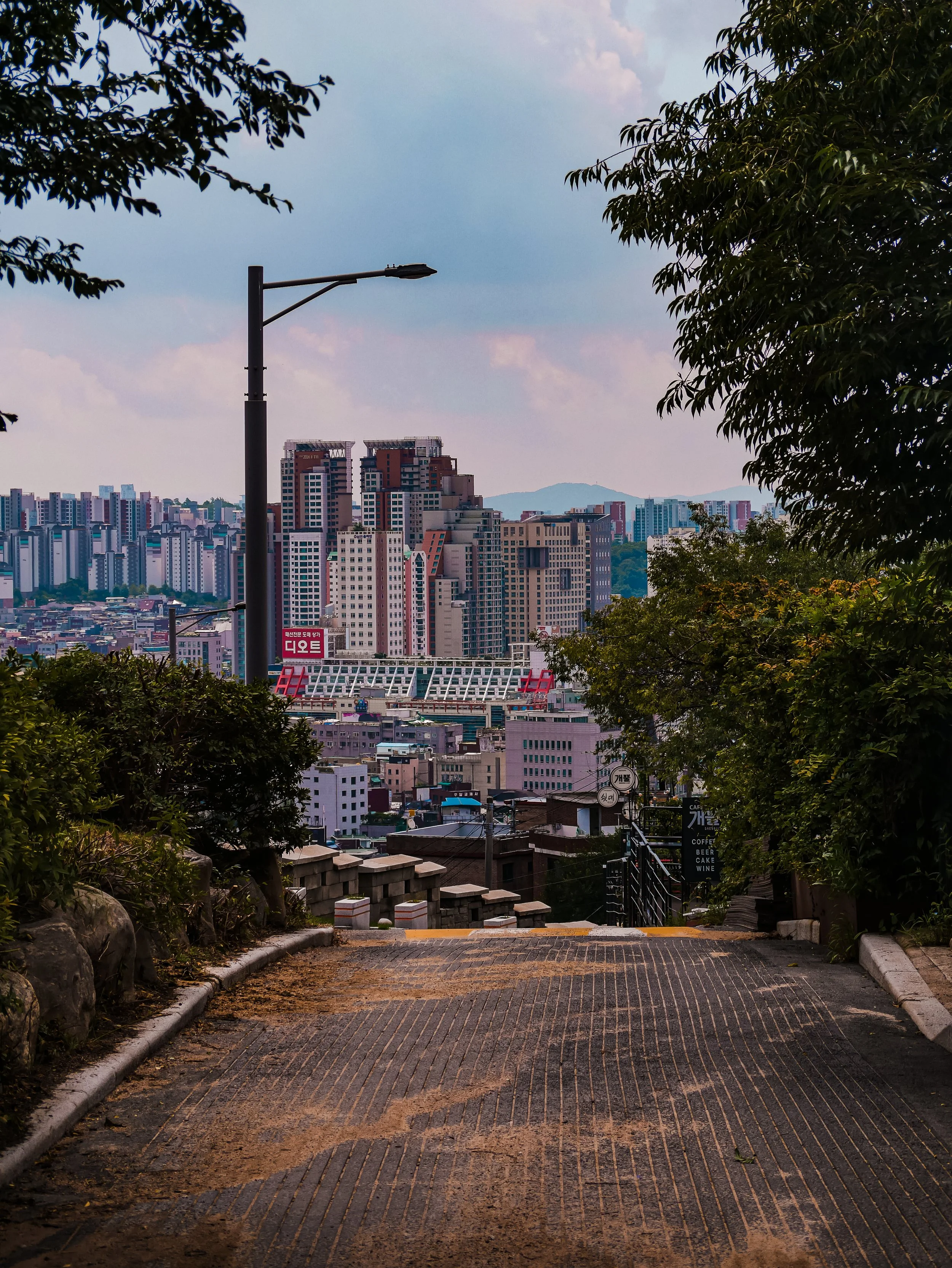 A cobblestone pathway descending between greenery on each side, leading towards a cityscape with tall buildings, a stadium, and a mountain in the background, under a partly cloudy sky.
