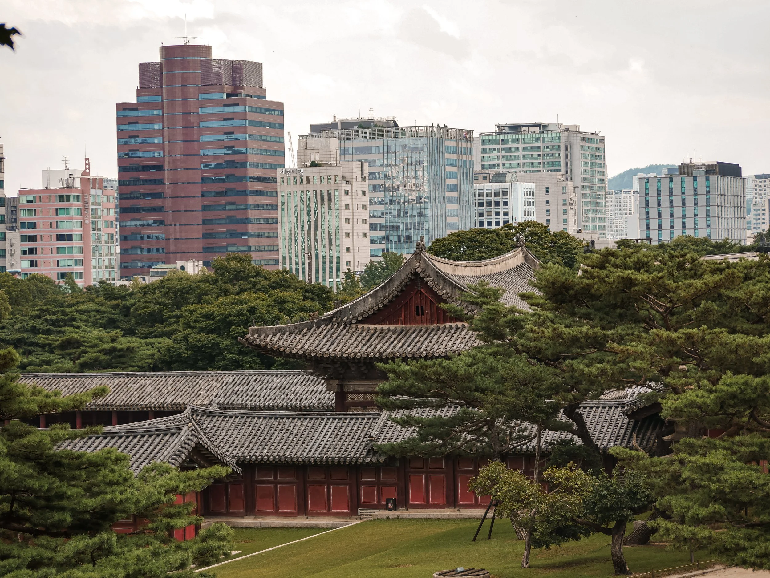 Traditional Korean temple with tiled roof and red wooden walls in the foreground, surrounded by lush green trees, with modern city buildings and skyscrapers in the background under a partly cloudy sky.