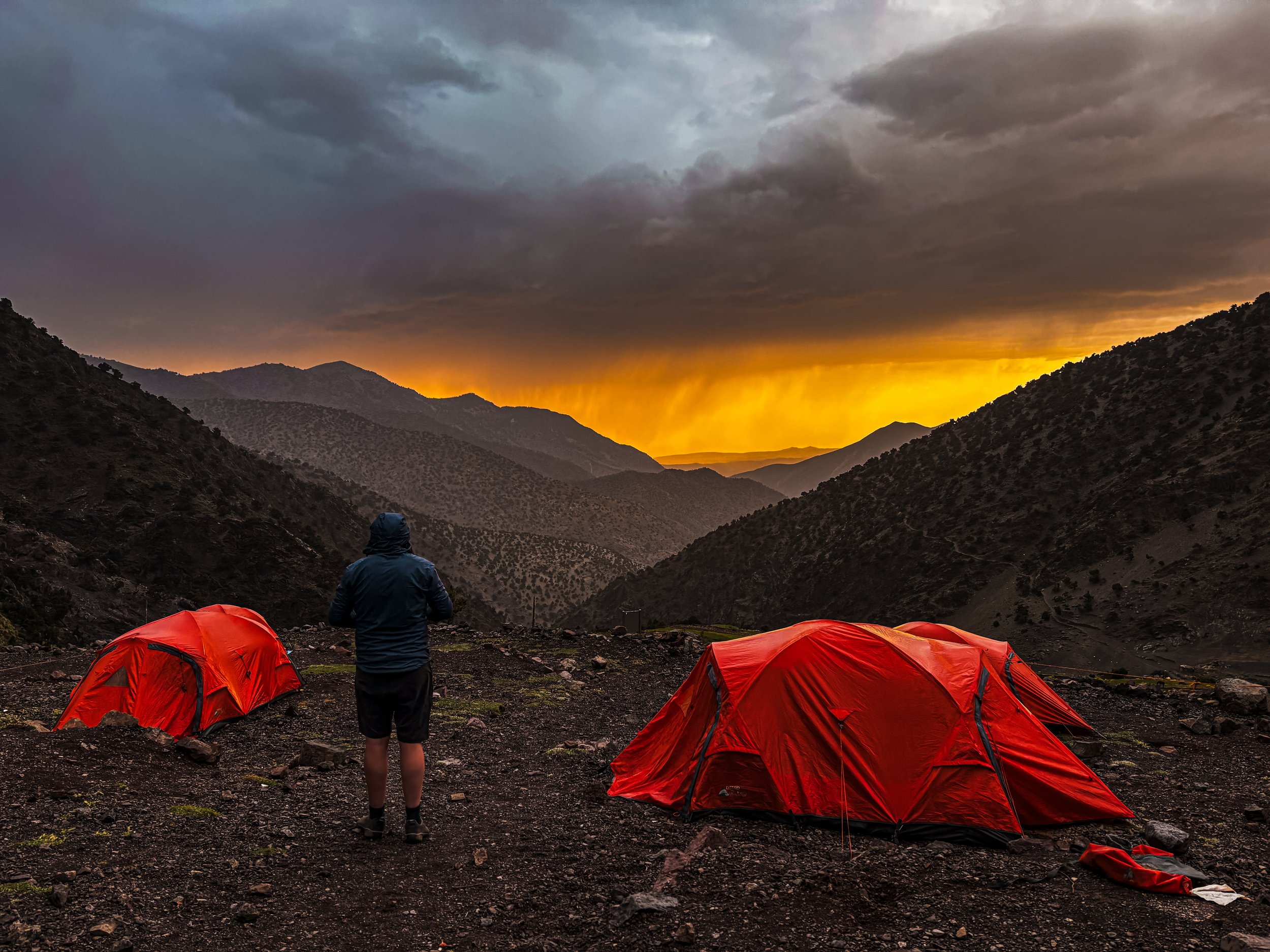 A person standing on rocky terrain in front of red tents in a mountain valley at sunset, with dark clouds overhead and a vibrant orange glow on the horizon.