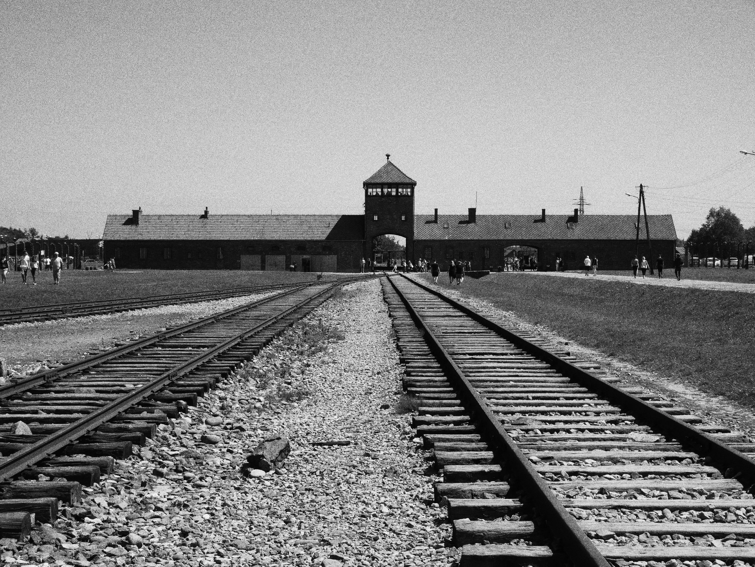Black and white photo of the Auschwitz concentration camp, showing railway tracks leading towards a large brick building with a central tower and an archway, surrounded by visitors.