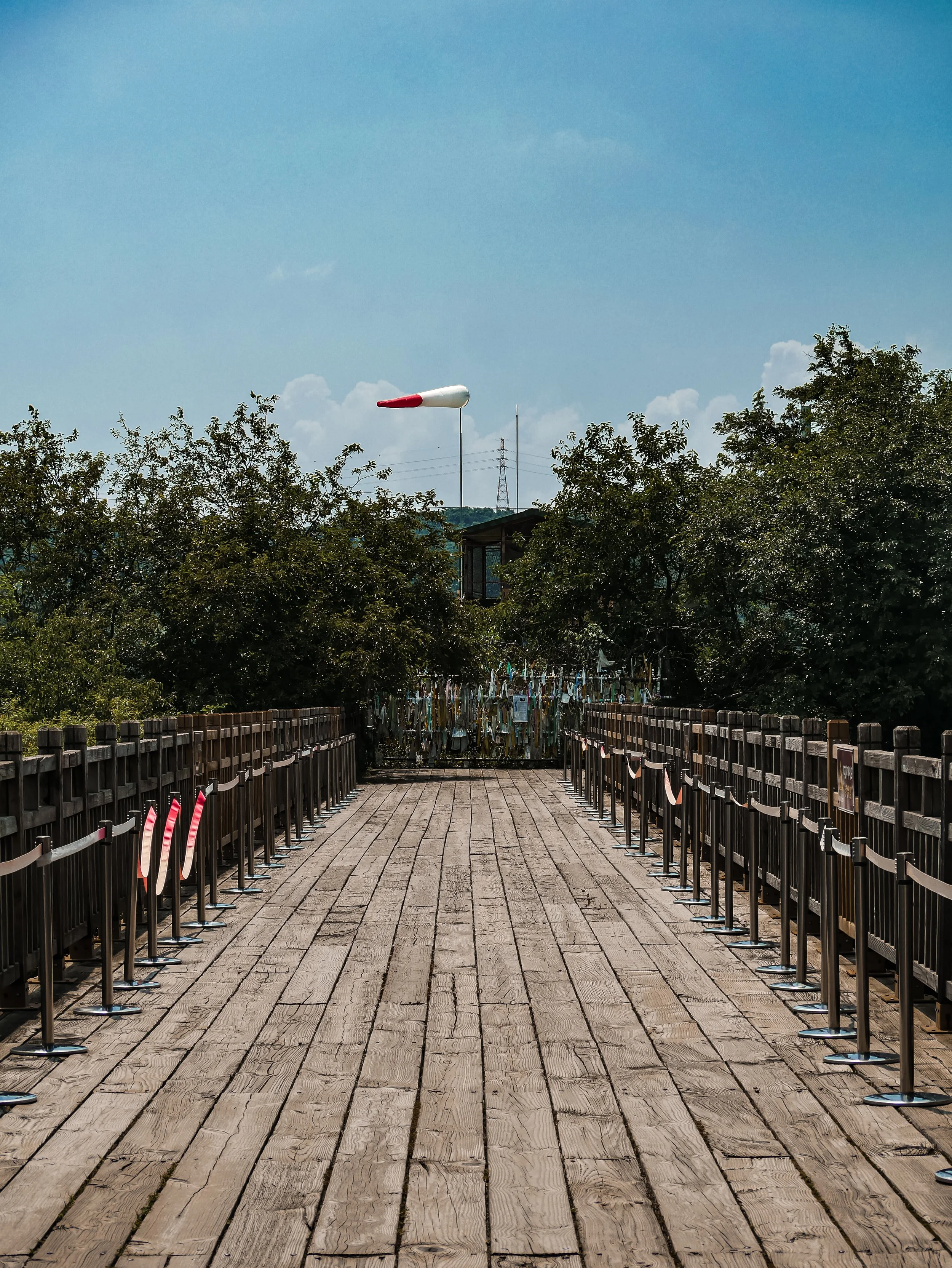 A wooden bridge with black railings leading to a garden area with laundry hanging, green trees, and a wind sock in the sky.