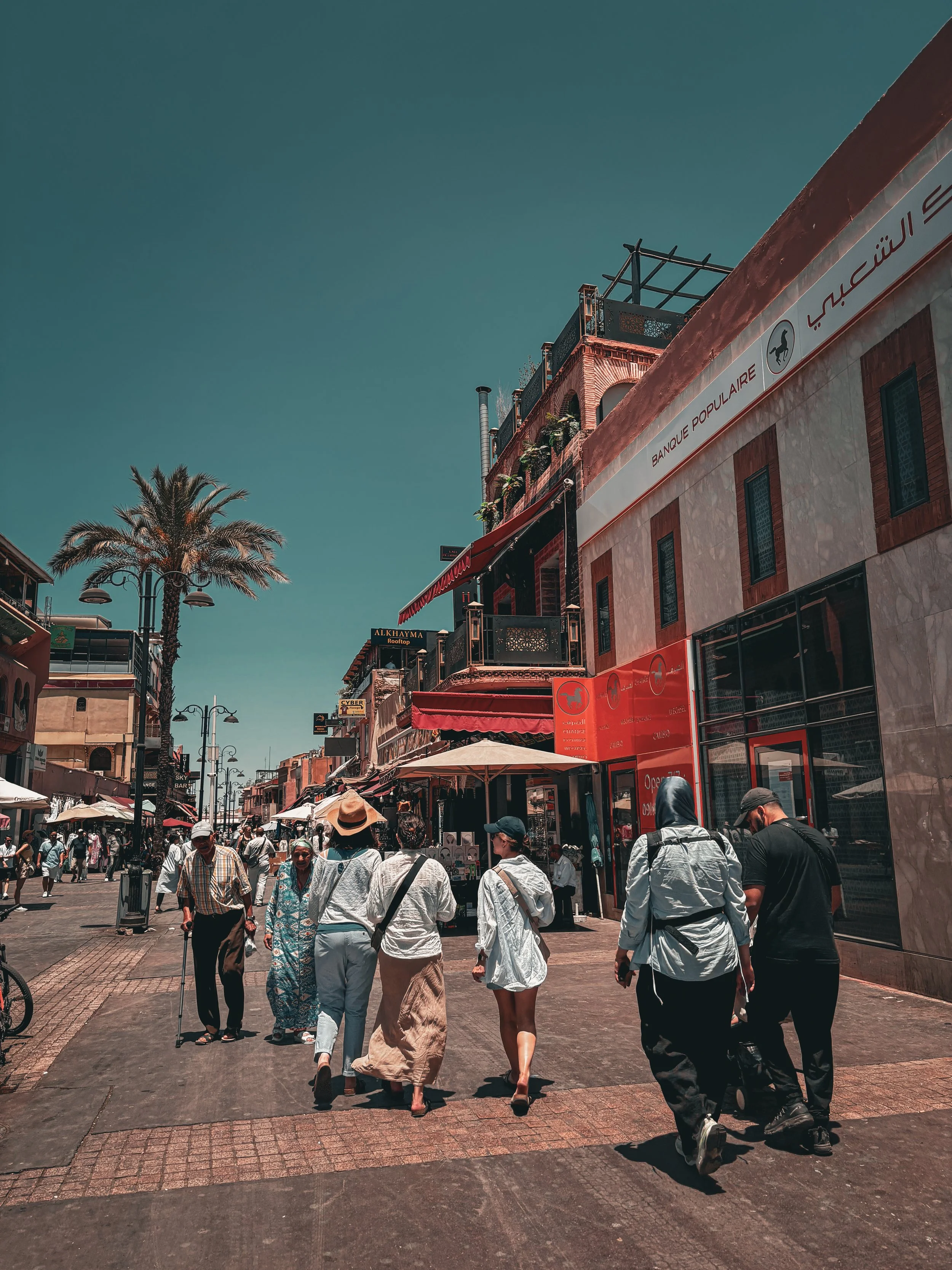 Street scene with people walking, shops, and colorful buildings, under a clear blue sky.
