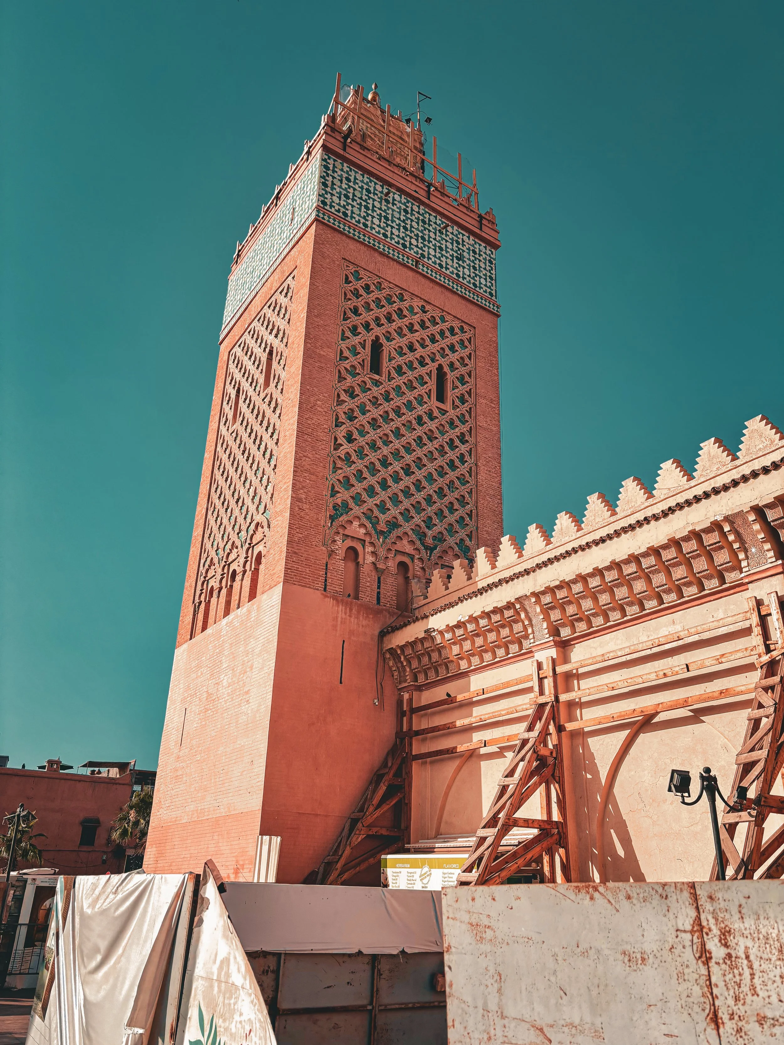 A tall, ornate pink-orange minaret with intricate geometric patterns and small arched windows against a clear blue sky, with construction scaffolding and barriers at its base.