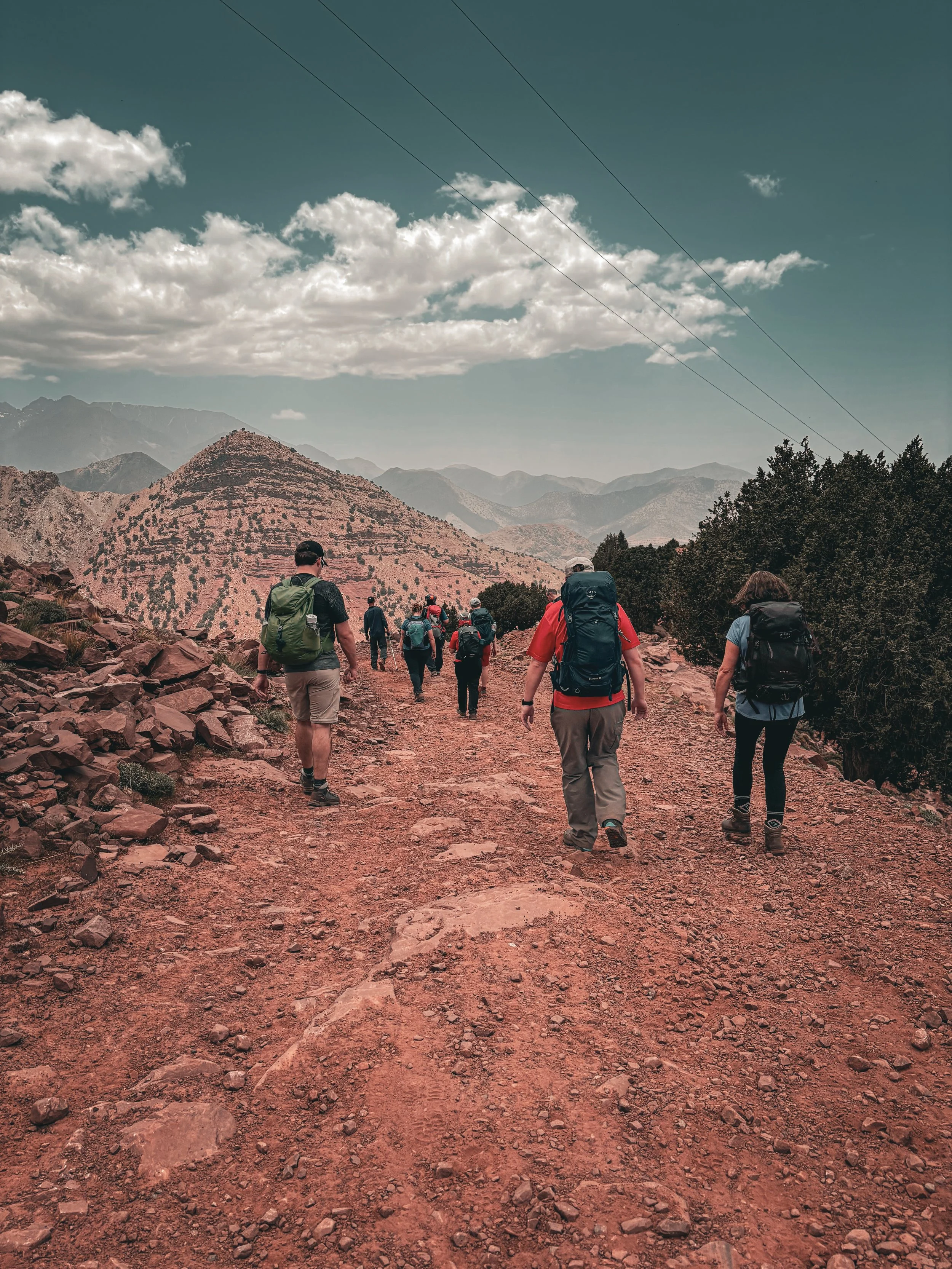 A group of people hiking on a rocky trail through a mountainous landscape with scattered trees, under a partly cloudy sky.