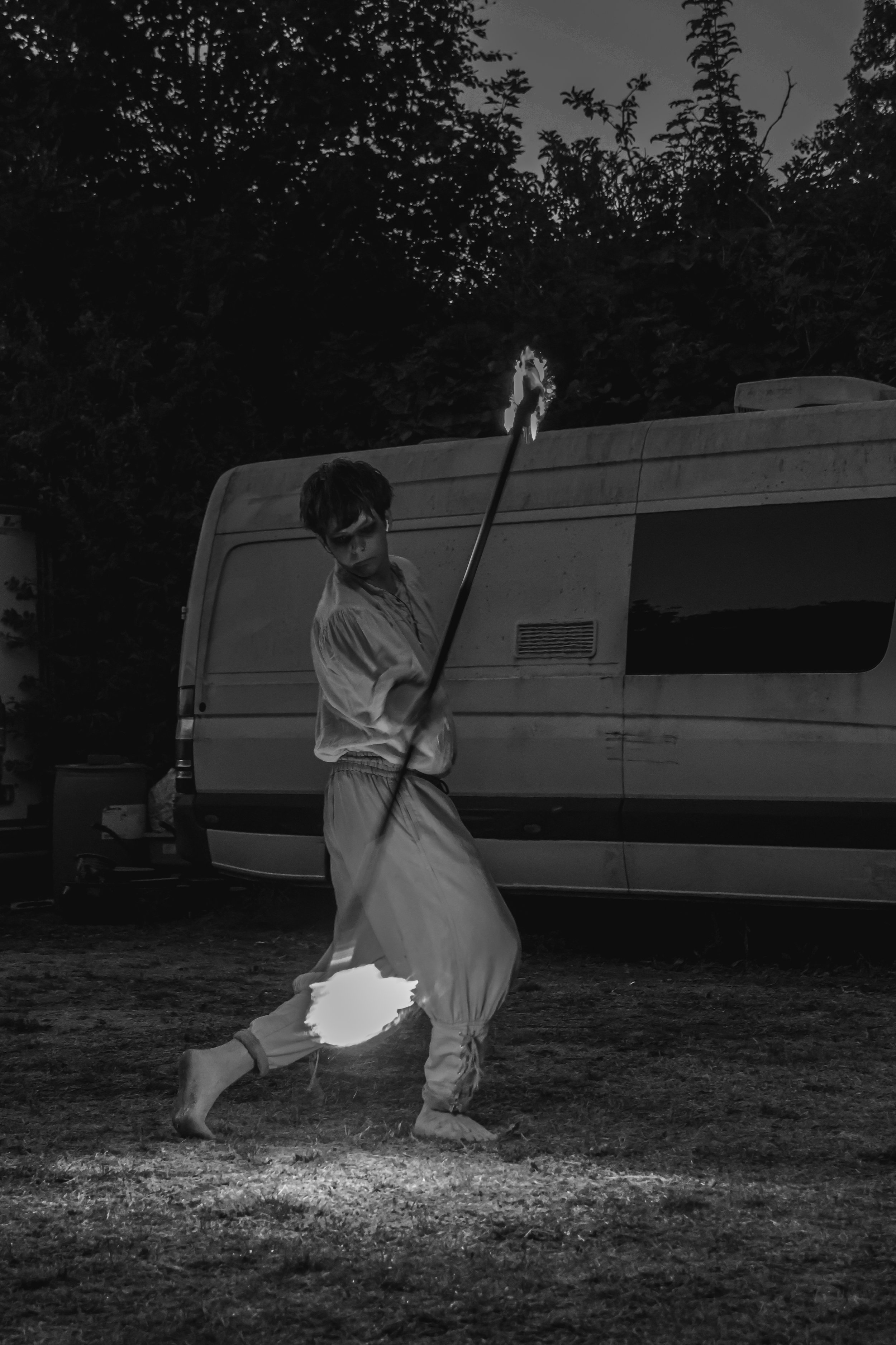 A person holding a flaming torch in front of a camper van at night, with trees in the background.