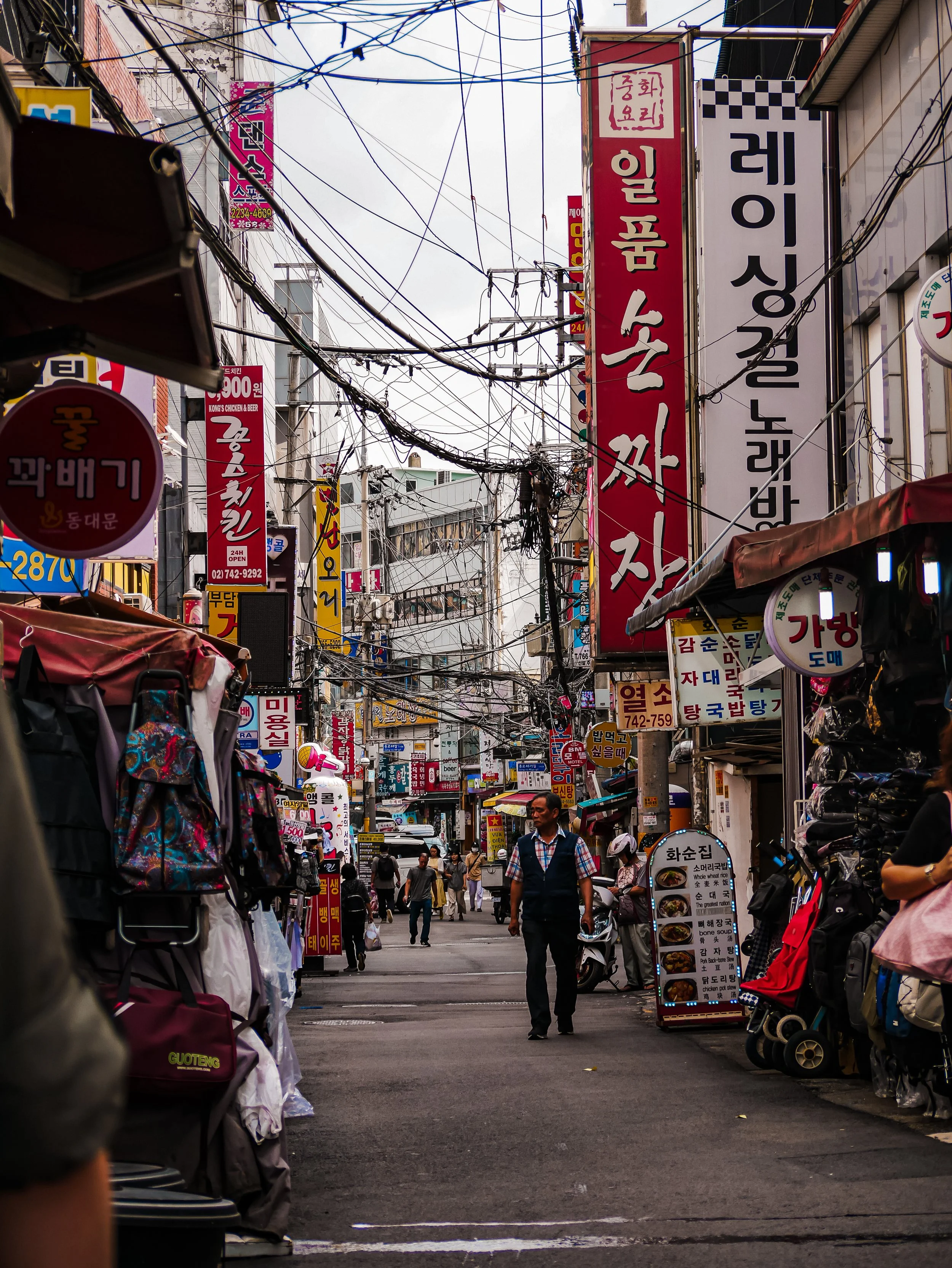 A busy street market with numerous signs in Korean, street vendors, and people walking through narrow alleyways, with overhead wires crisscrossing above.