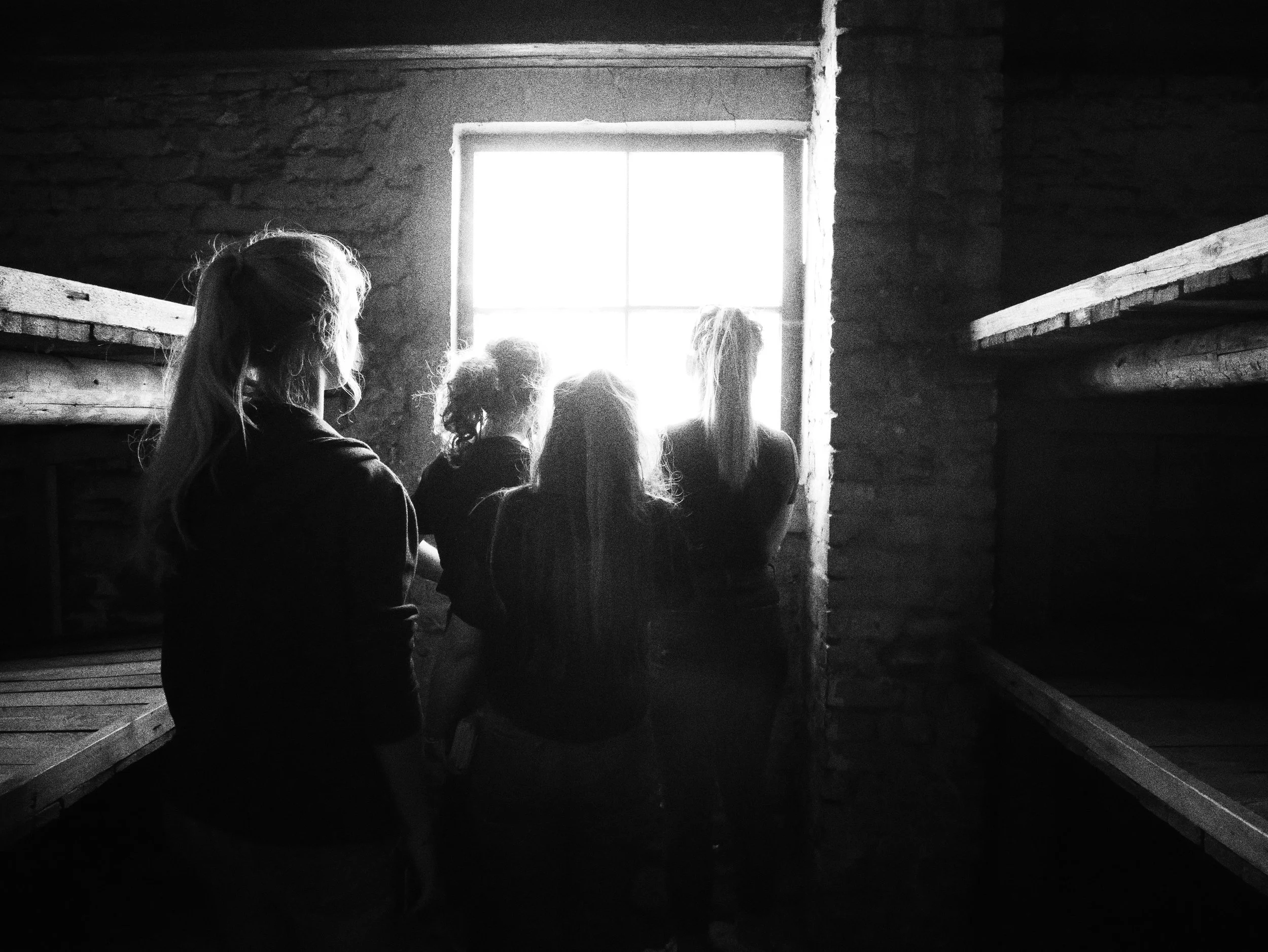 Black and white photo of four people sitting inside a rustic cabin, looking out a window with bright light shining through, with wooden beams and walls.