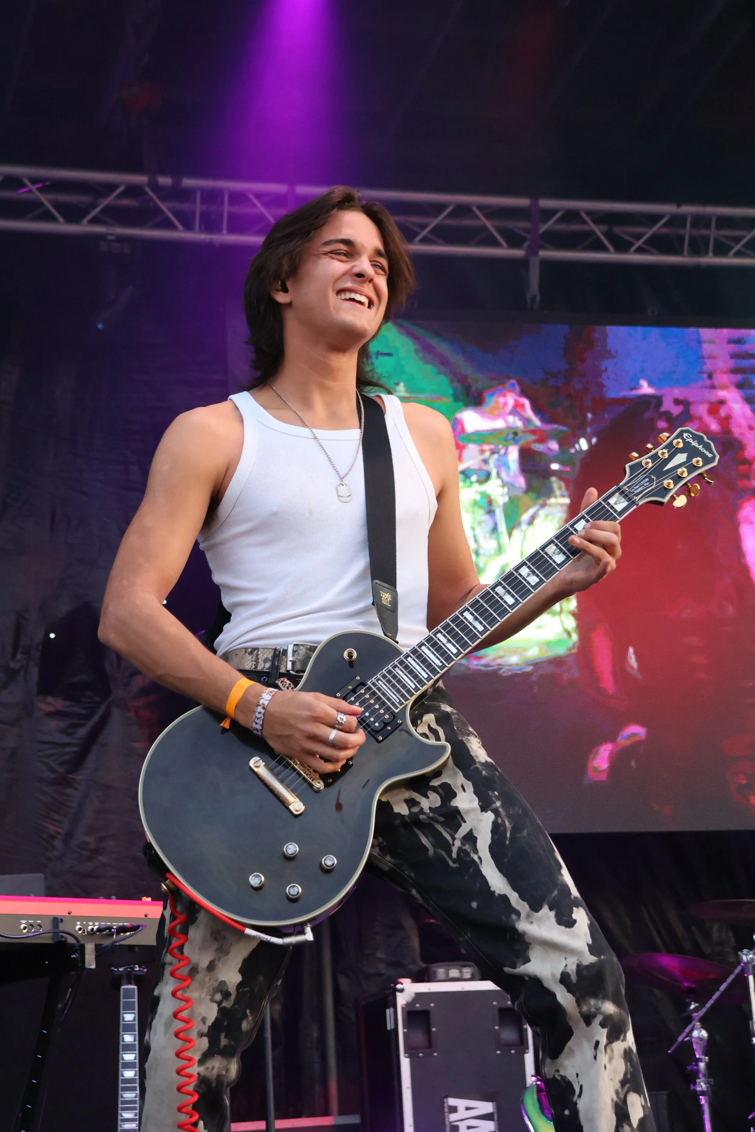 A young man playing an electric guitar on stage during a live performance, smiling with purple and pink stage lighting behind him.