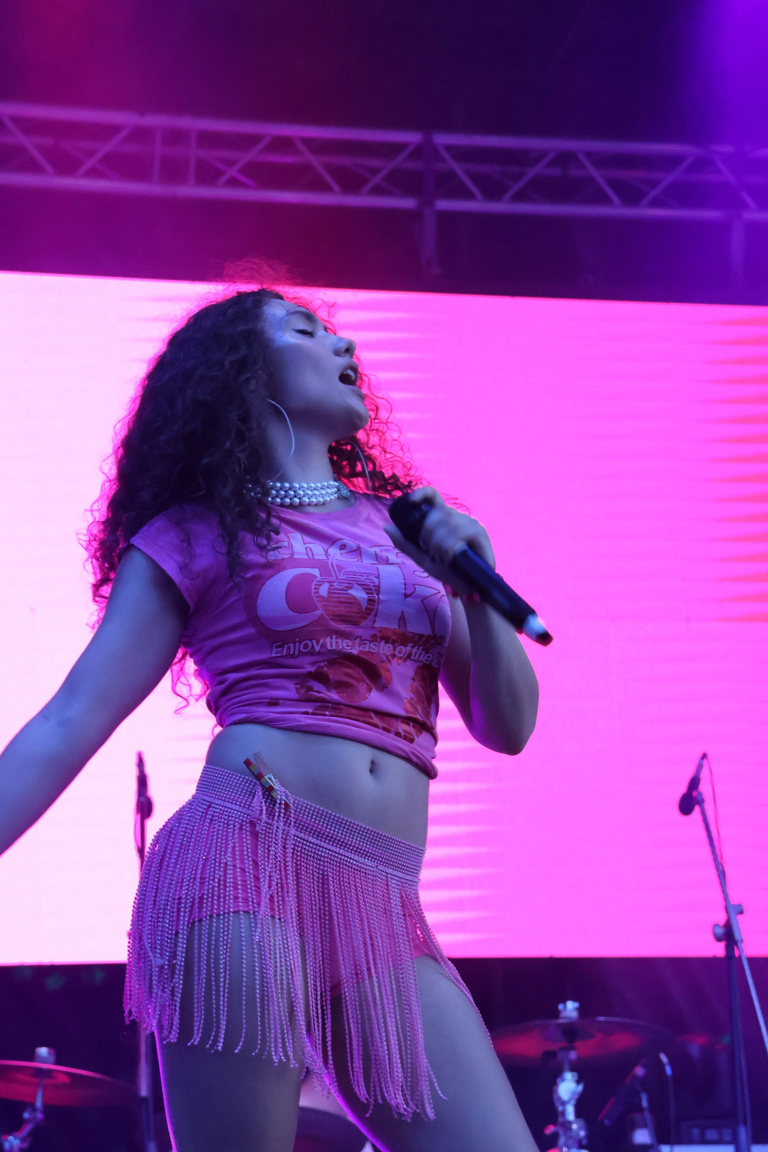 A woman singing on stage holding a microphone, with a pink background, wearing a pink T-shirt, pearl necklace, and a pink beaded skirt.