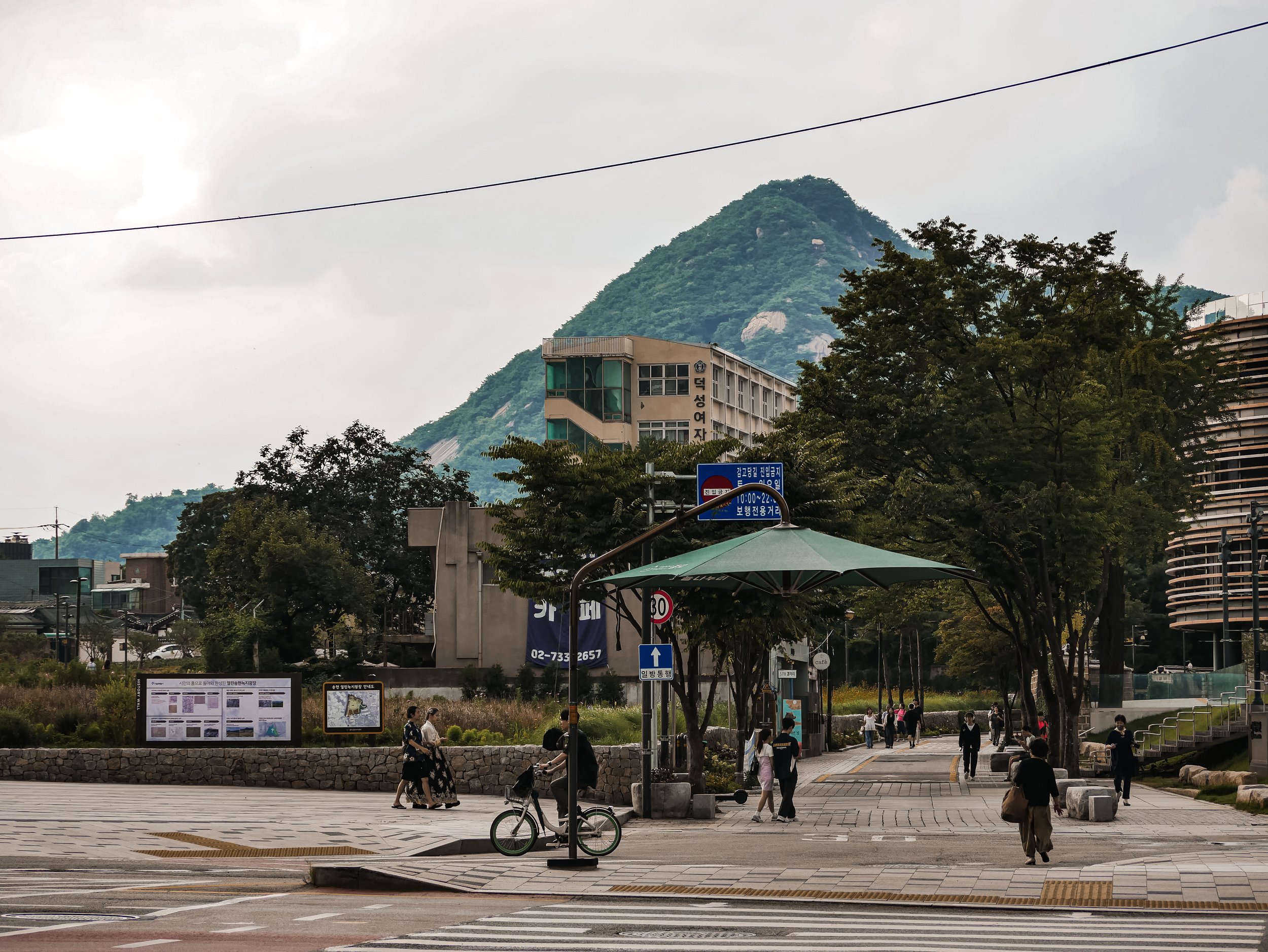 A busy urban area with pedestrians walking, some on a crosswalk, a cyclist, and a building with a mountain in the background. The scene includes trees, signage, and a cloudy sky.