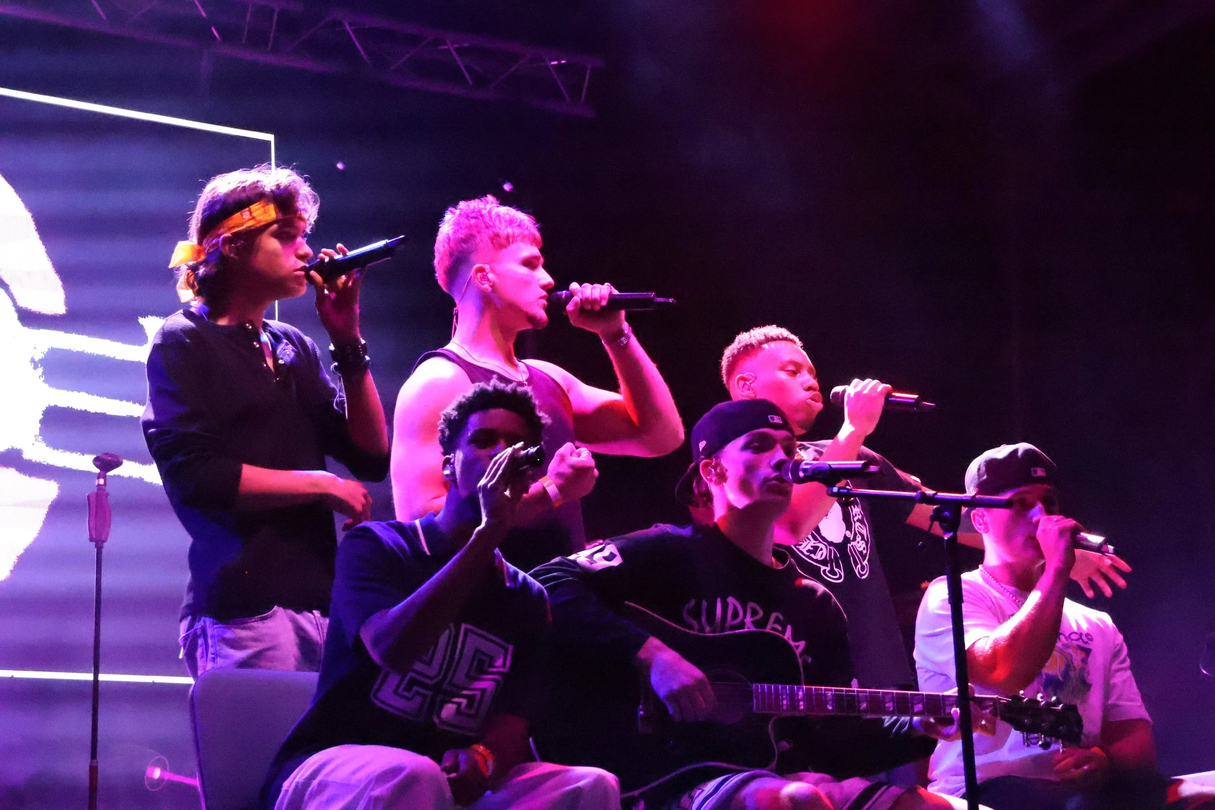 Group of young people on stage, some singing into microphones and one playing guitar, under purple stage lighting.