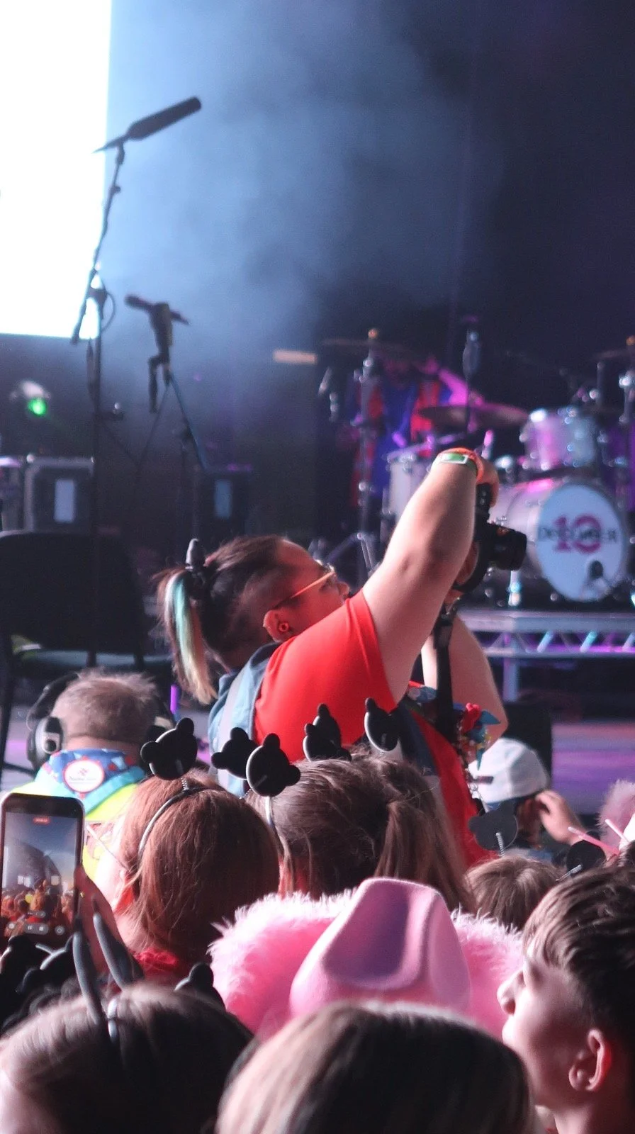 Crowd of children and adults at a concert or performance, with some wearing Mickey Mouse ears, taking photos, and facing a stage with musical instruments and performers.