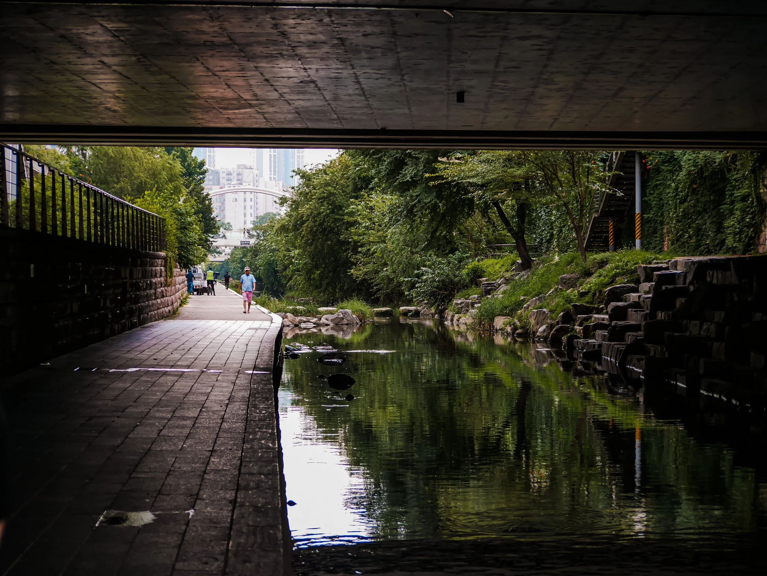 View of a city park from underneath a bridge, showing pedestrians walking alongside a small stream, with lush green trees and buildings in the background.
