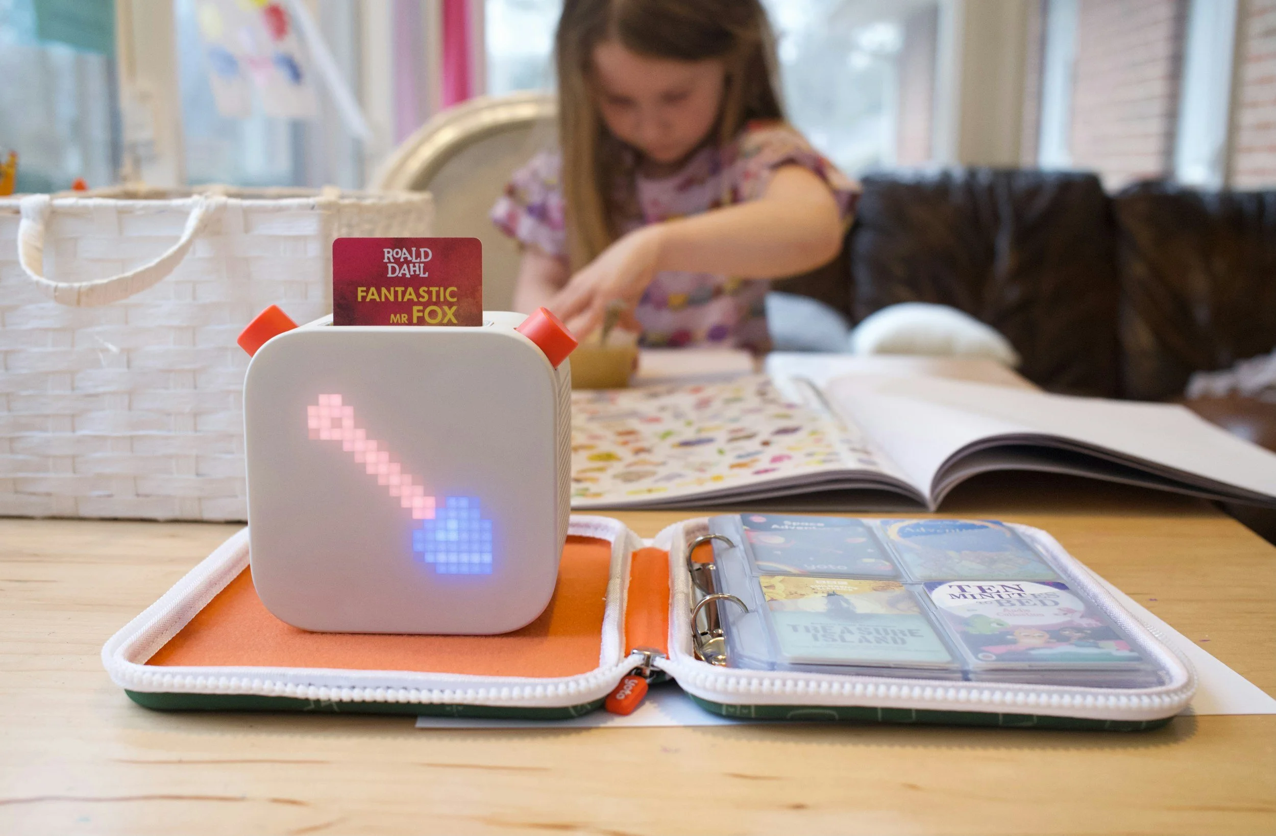 A small robot speaker with a pixelated heart display on its front, placed on a gift box. A girl in the background is engaged in an activity at a table, with books and a black dog nearby, in a room with windows and bright light.
