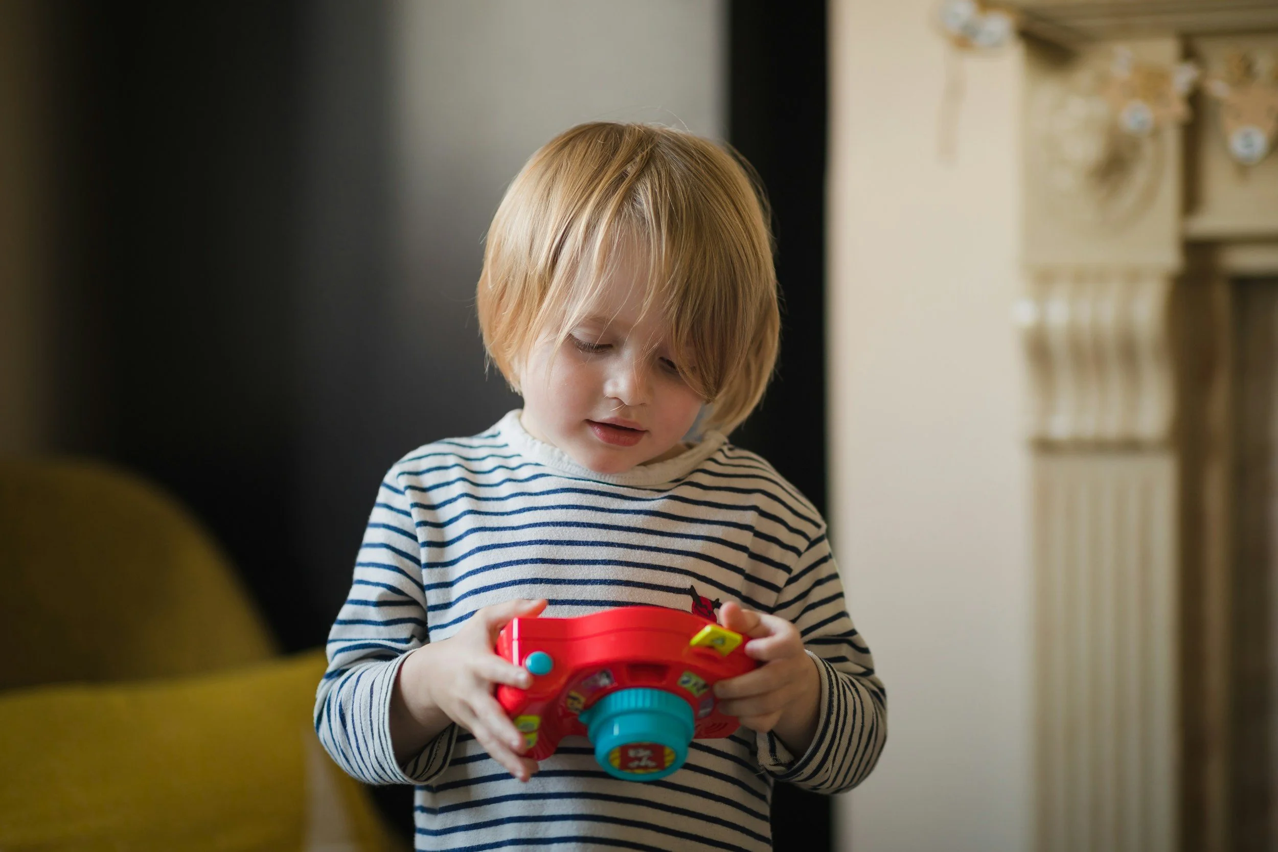 A young child with blonde hair wearing a striped shirt is focusing on a colorful toy or device in her hands.
