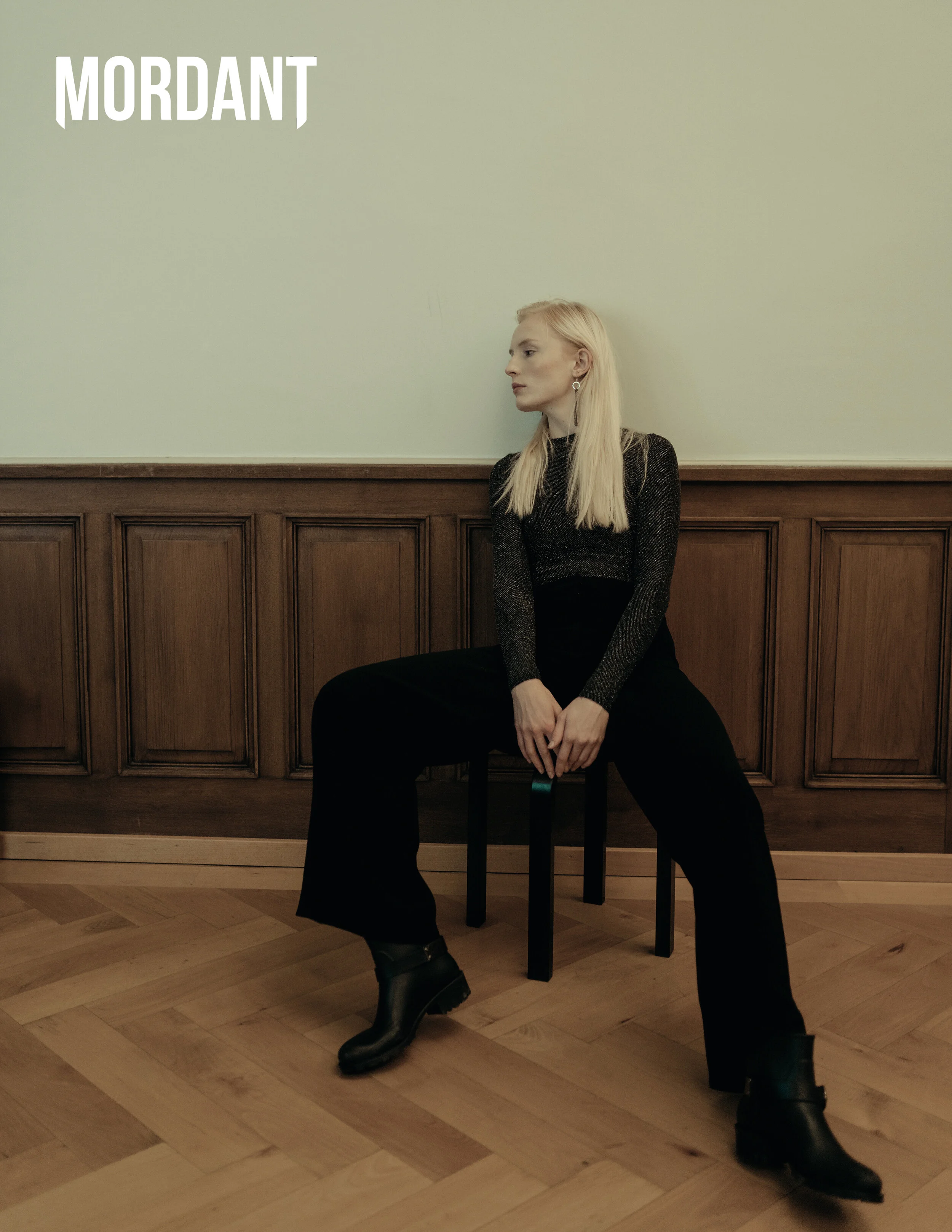 A woman with long blonde hair sitting on a black stool in front of a light-colored wall with wooden paneling, wearing a black long-sleeve top, black wide-leg pants, and black boots.