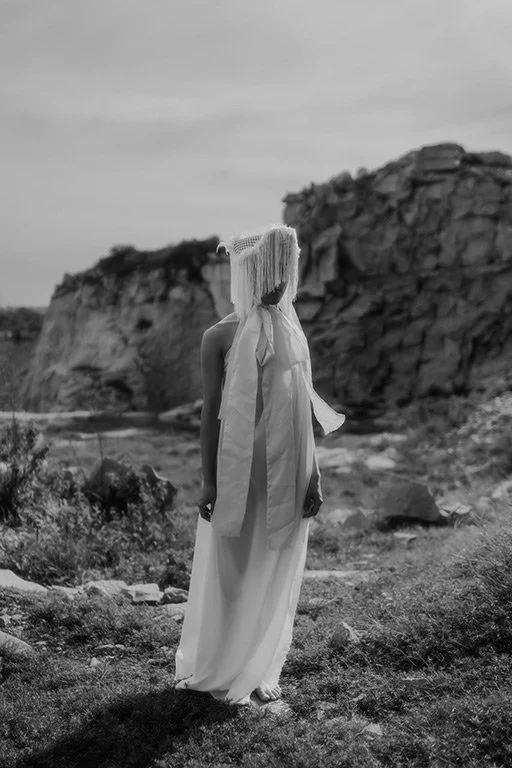 A person standing outdoors near rocky terrain, wearing a long dress, a wide-brimmed sun hat, and a light scarf around their neck, with their face turned away from the camera.