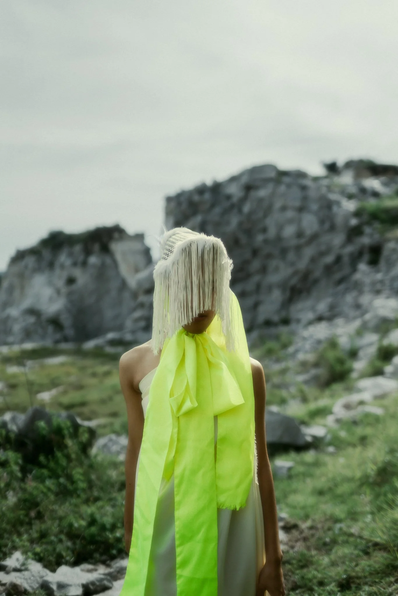A person in a white dress with a large, neon yellow-green bow and matching scarf stands outdoors near rocky hills and green vegetation.