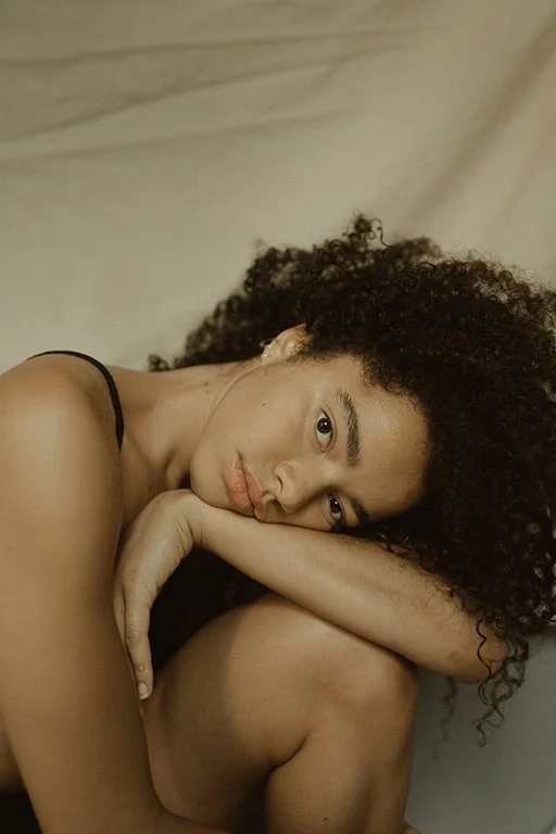A woman with curly hair lying on her side with her head resting on her knee, looking at the camera with a thoughtful expression.