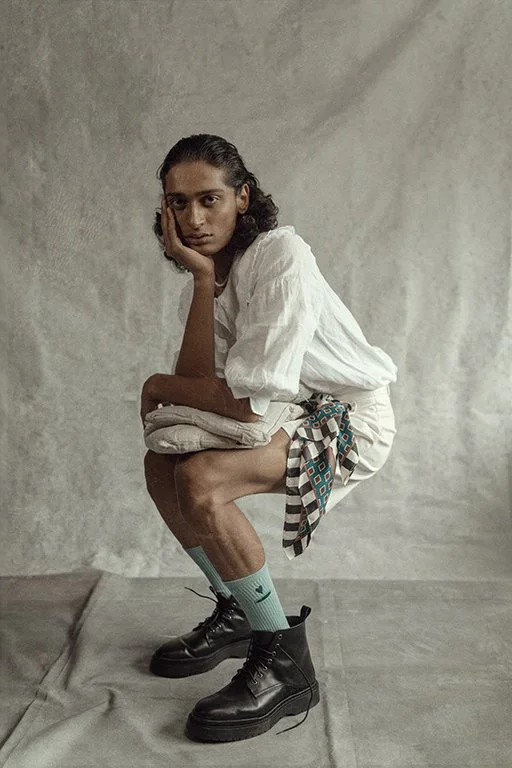 A woman in casual clothing, with a white shirt and patterned shorts, is squatting on the floor against a plain backdrop, resting her head on her hand and looking at the camera.
