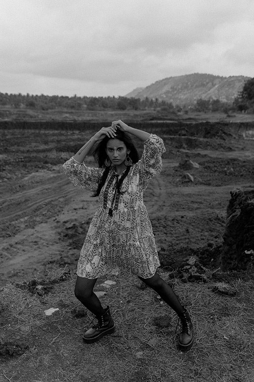 A woman in a patterned dress and combat boots standing on a dirt path in an open landscape with hills in the background, black and white photo.