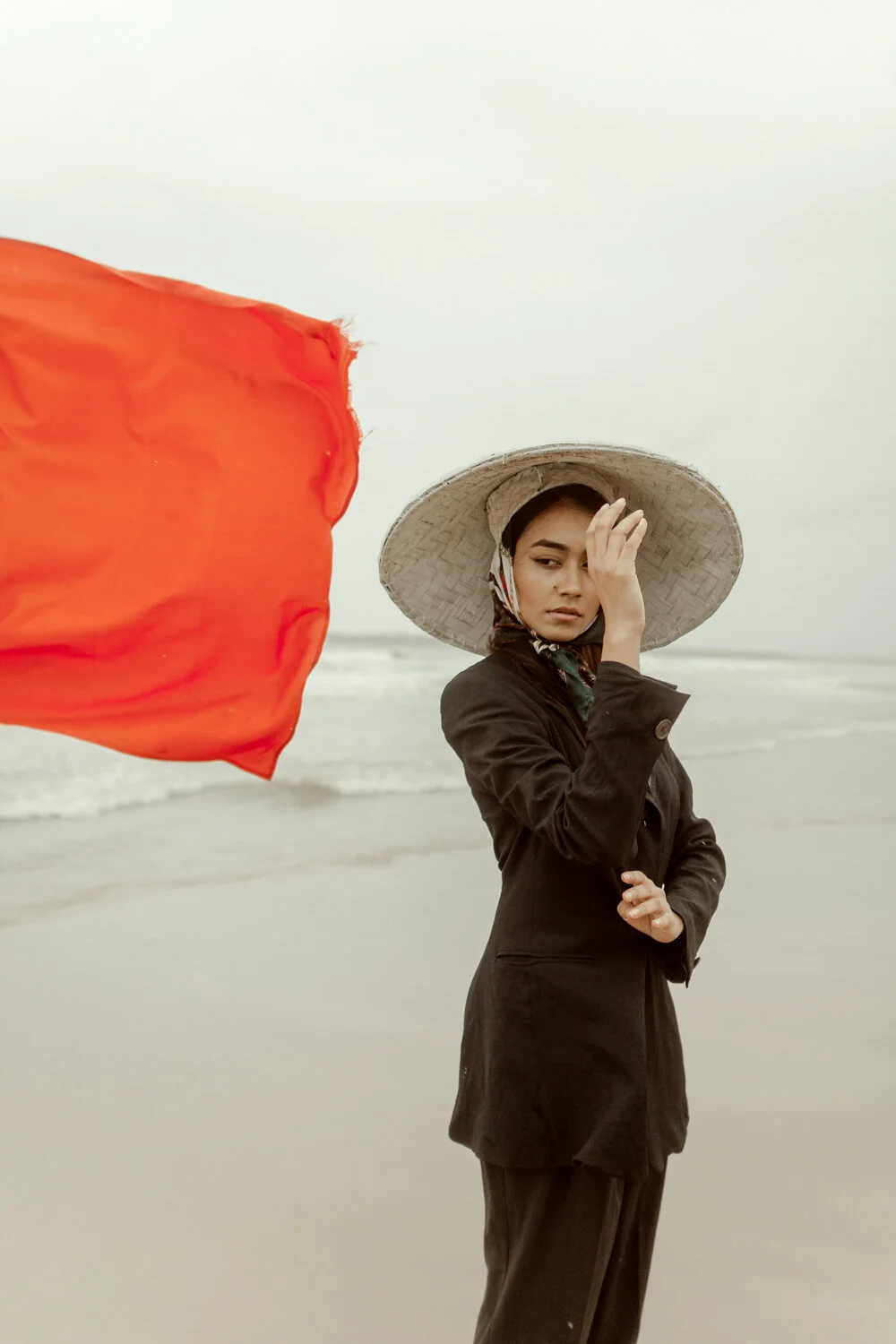 A woman in a black coat and large straw hat standing on a beach with a red flag blowing in the wind, overcast sky.