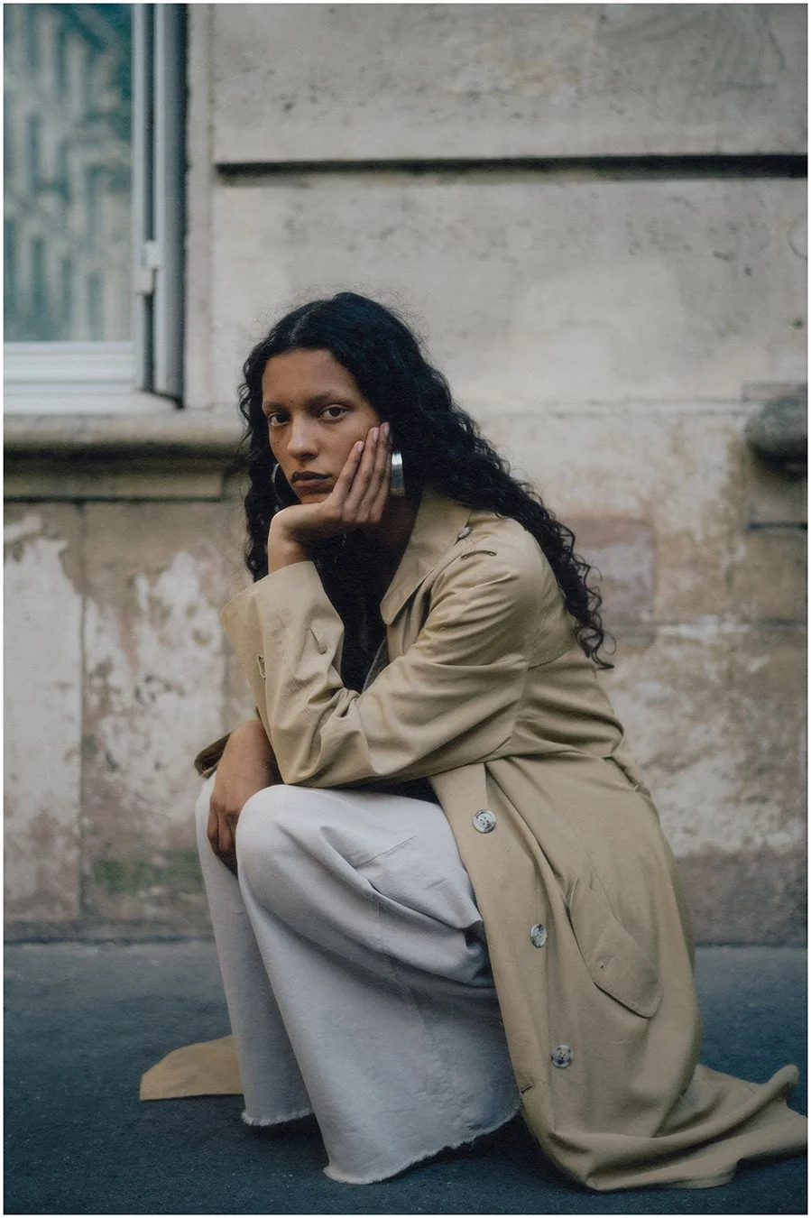 A woman with long curly black hair, wearing a beige trench coat and white pants, squatting on the ground with her hand resting on her face, in front of a worn concrete wall.