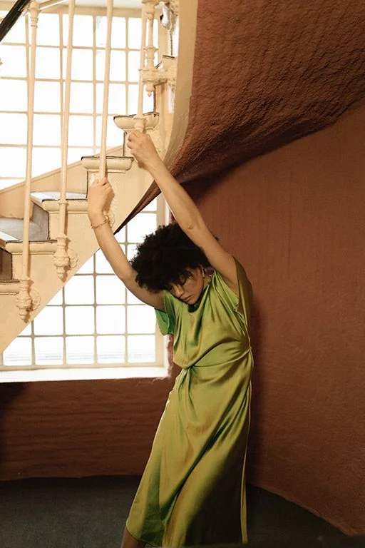 A woman in a green satin dress stands under a staircase, leaning against a textured orange wall, with one arm reaching up to touch the staircase railing.