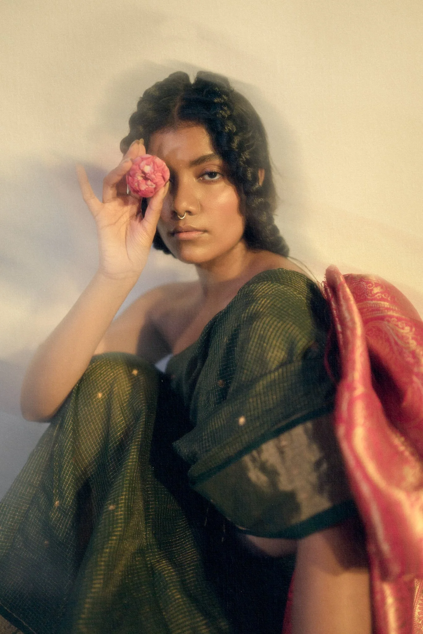 A woman with curly hair and a septum piercing holding a pink flower to her eye, sitting with a draped patterned fabric over her shoulder.