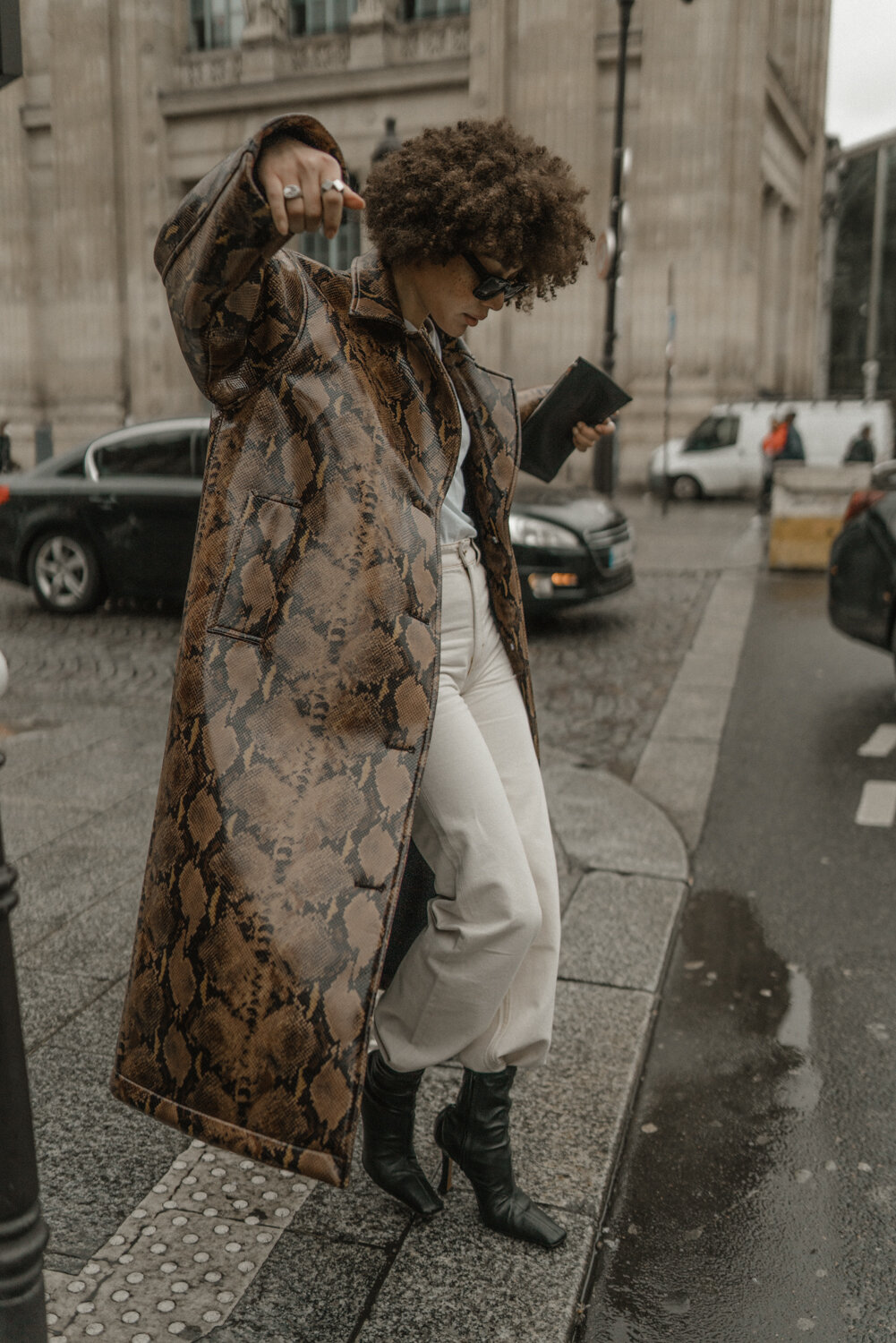 A woman with curly hair, wearing sunglasses, a long snake print coat, white pants, and black high-heeled boots, stands on a city sidewalk checking her phone.