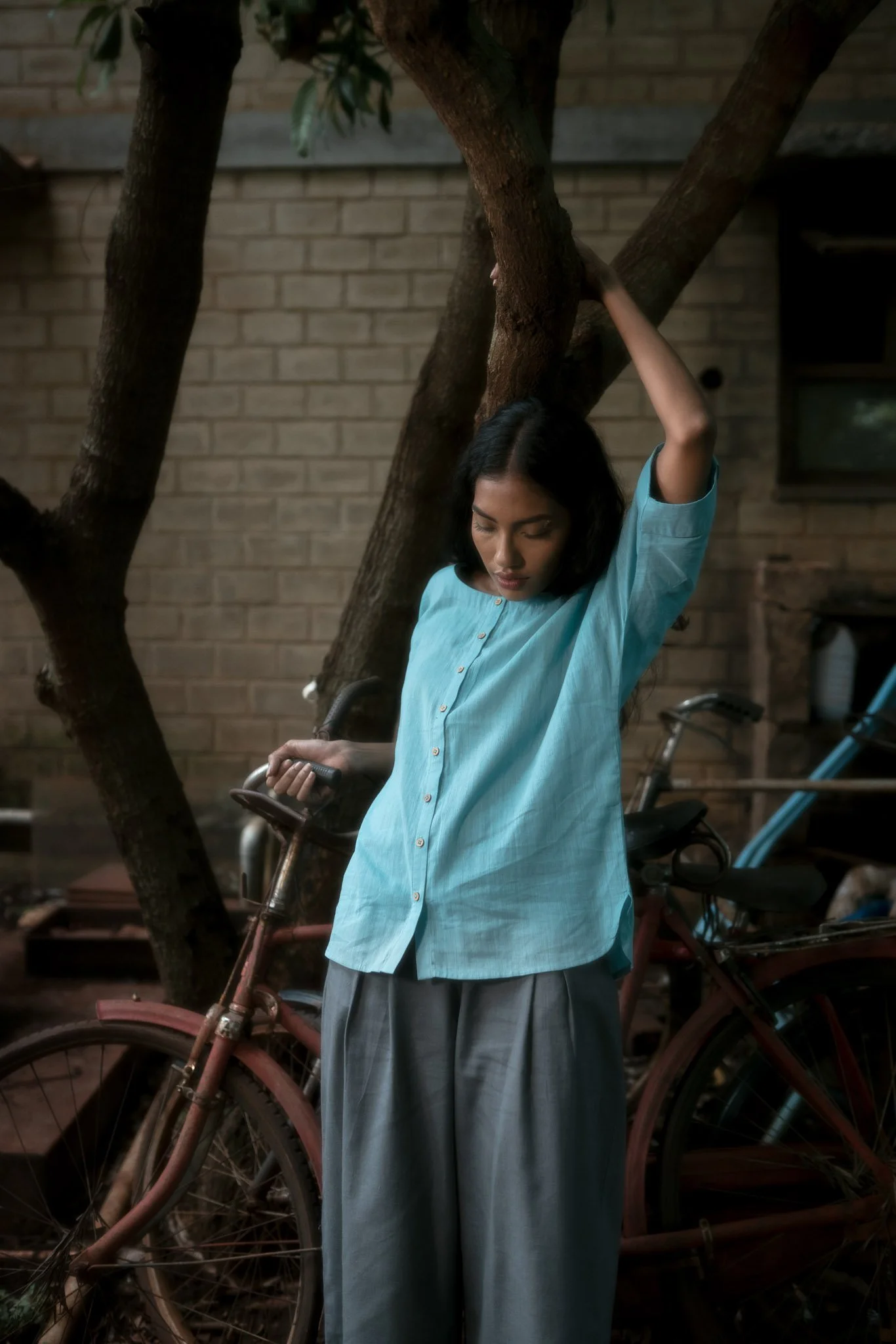 A woman in a blue shirt and gray pants stands outdoors next to a tree, holding the tree with one hand raised above her head and the other hand on a bicycle handlebar, with bicycles and a brick wall in the background.