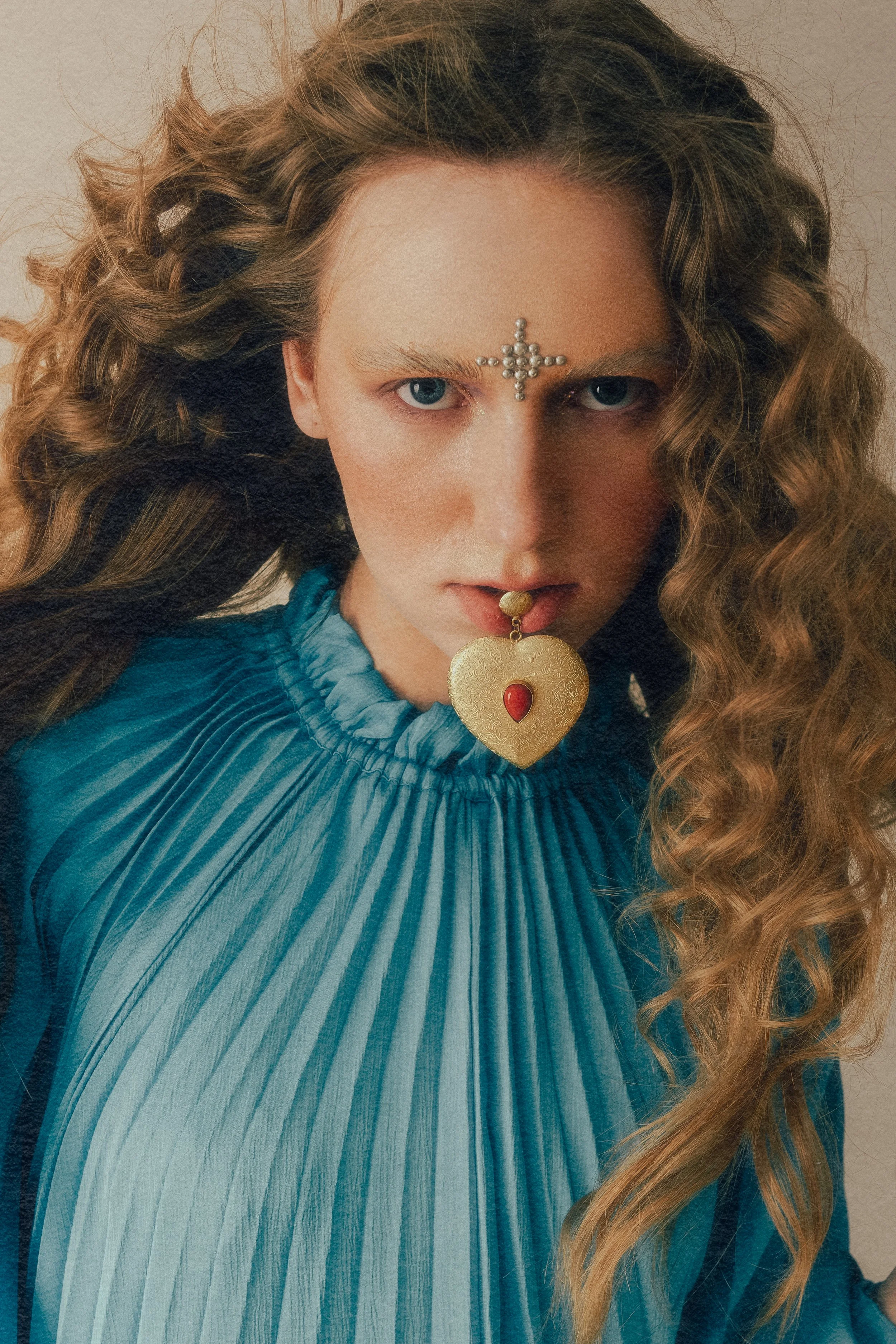 Close-up of a woman with curly hair, blue eyes, and light skin wearing a blue pleated top, with decorative face jewelry including a cross on her forehead and a heart-shaped pendant in her mouth.