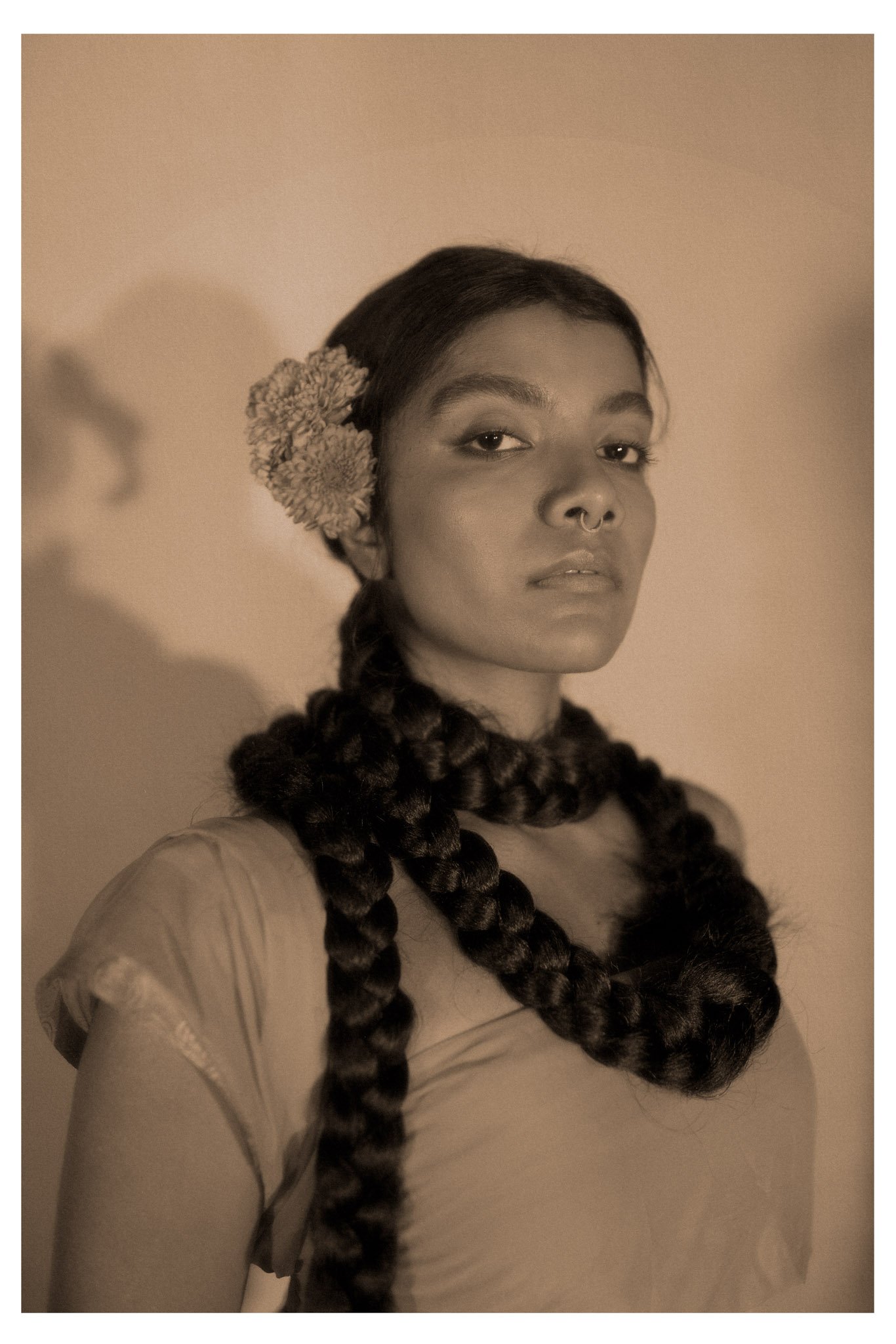 A woman with long, thick, braided hair wearing a flower hair accessory, a septum piercing, and a casual top against a simple background.