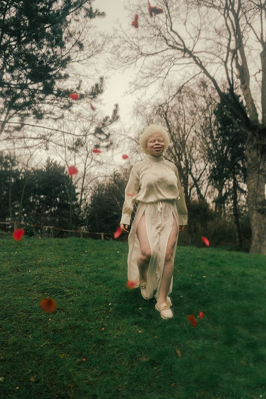 A woman with white curly hair walking on grass in a park with falling red flower petals, surrounded by leafless trees.