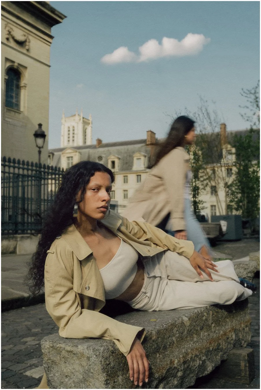 A woman with long dark curly hair lying on a stone bench outdoors in a cityscape, with historic-style buildings and a tower in the background. She is wearing a tan jacket and white clothing and looking directly at the camera.