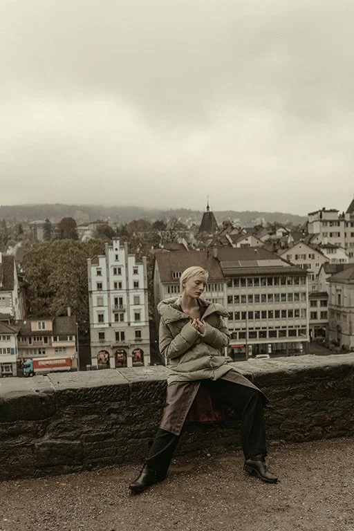A woman sitting on a stone ledge overlooking a cityscape with various buildings, on an overcast day.