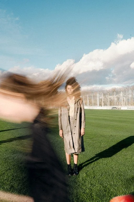A woman standing on a grassy field with a cloudy sky and some sailboat masts in the background, while someone with long hair moves quickly past her, creating a blurred effect.