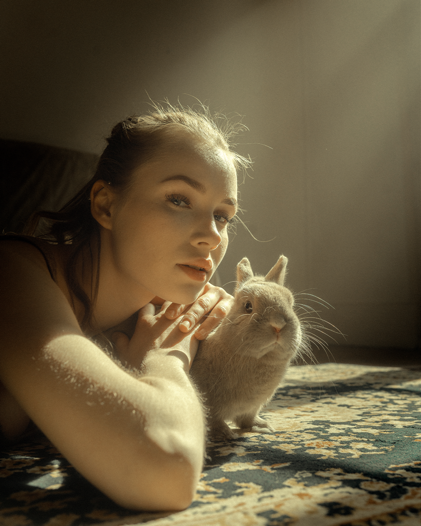 A young woman with light skin, long hair, and makeup, lying on her stomach on a patterned rug, resting her head on her arm, next to a small, light-colored rabbit.
