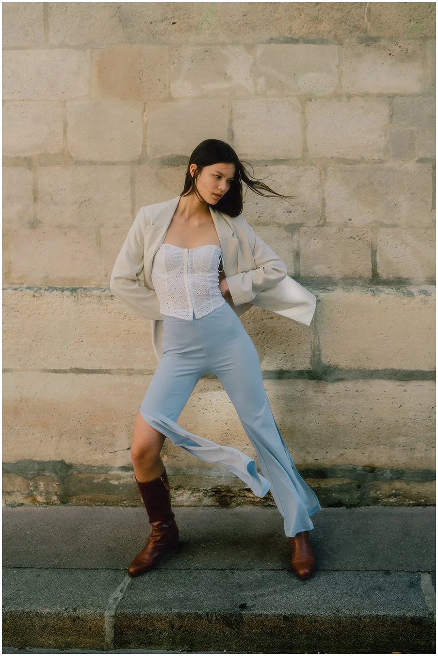 A woman with dark hair wearing a white corset top, light blue wide-leg pants with a slit, a beige blazer, and brown boots, standing in front of a beige stone wall with her hands on her hips.