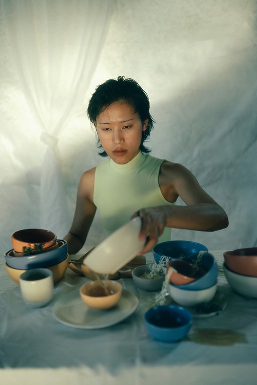 A woman with short dark hair wearing a sleeveless yellow top is pouring water into bowls on a table with various dishes and cups, in a softly-lit setting with a cream-colored curtain in the background.
