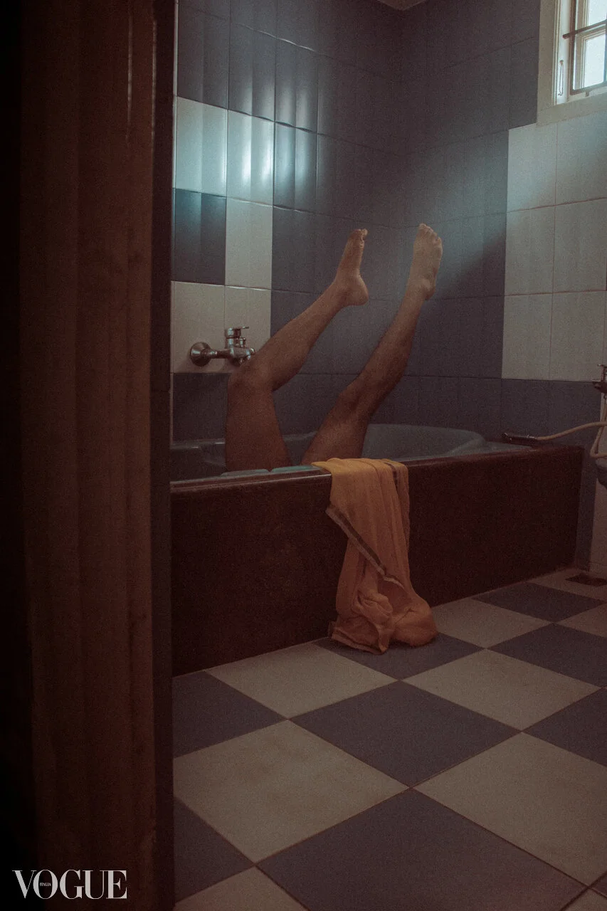 Person with legs raised in a bathtub, with a towel hanging over the edge, in a bathroom with checkered floor tiles and tiled walls.