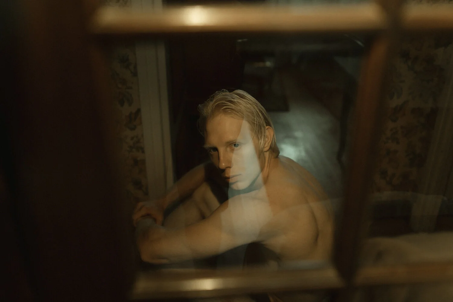 Looking down through a glass window at a young man with blond hair, sitting on the floor with his arm resting on his knee, in a dimly lit room with patterned wallpaper.