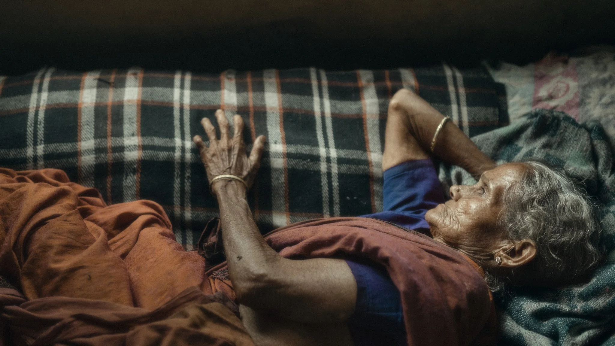 An elderly woman with gray hair and wearing jewelry, lying on a bed with a checkered blanket, resting her head on a pillow, and wearing a multicolored outfit.