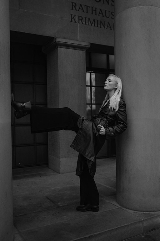 A woman leaning against a column outside a building with the words 'Rathaus Kriminal' visible, wearing a long coat, holding a smaller coat, with one leg raised resting against the wall.