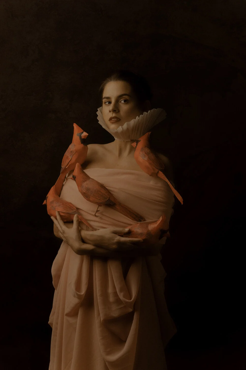 A woman with dark hair and light makeup wearing a nude-colored dress with a ruffled collar, surrounded by orange birds, against a dark background.