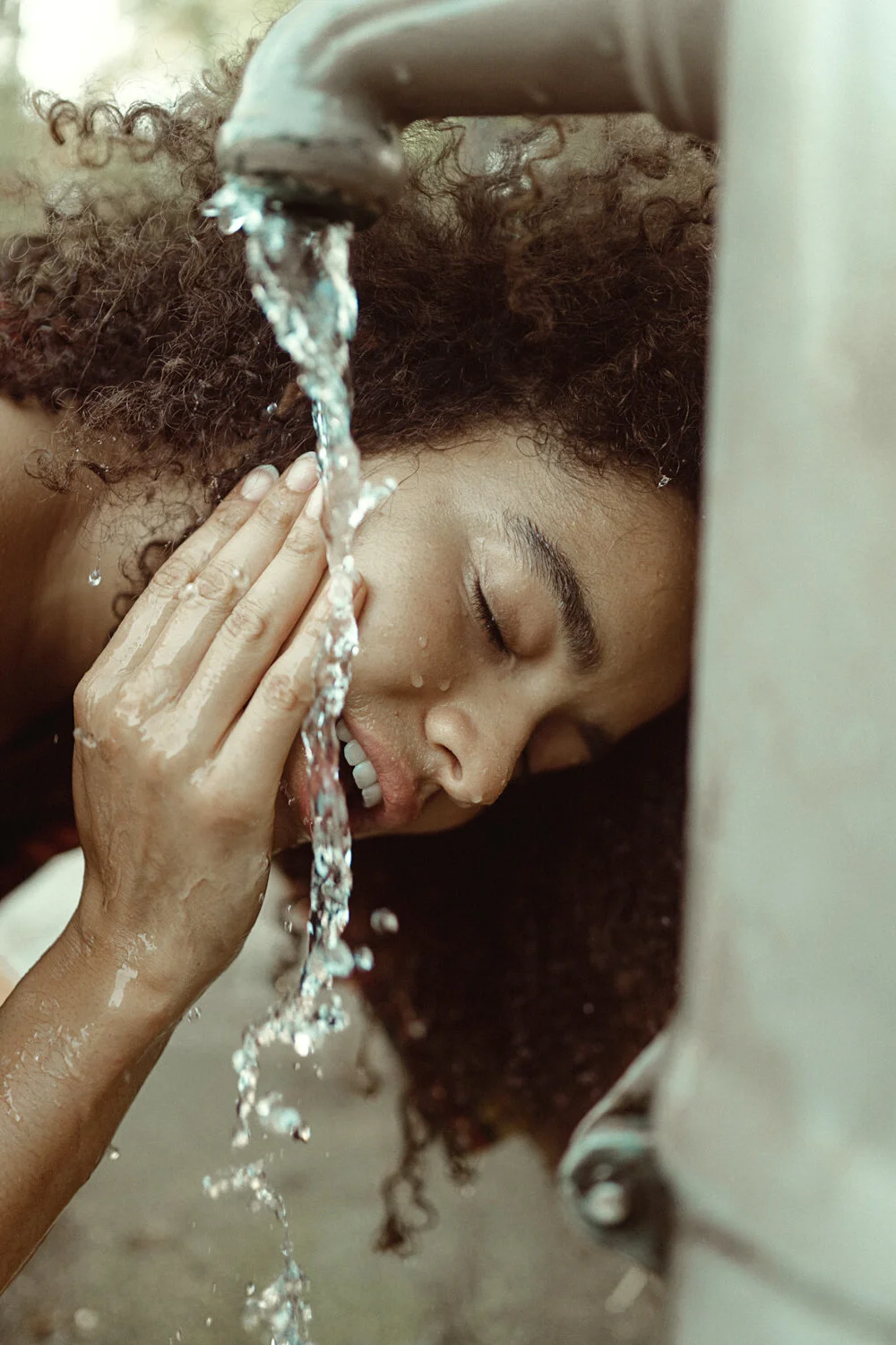A woman with curly hair leaning under a running sink faucet with water, eyes closed, touching her cheek with her hand.